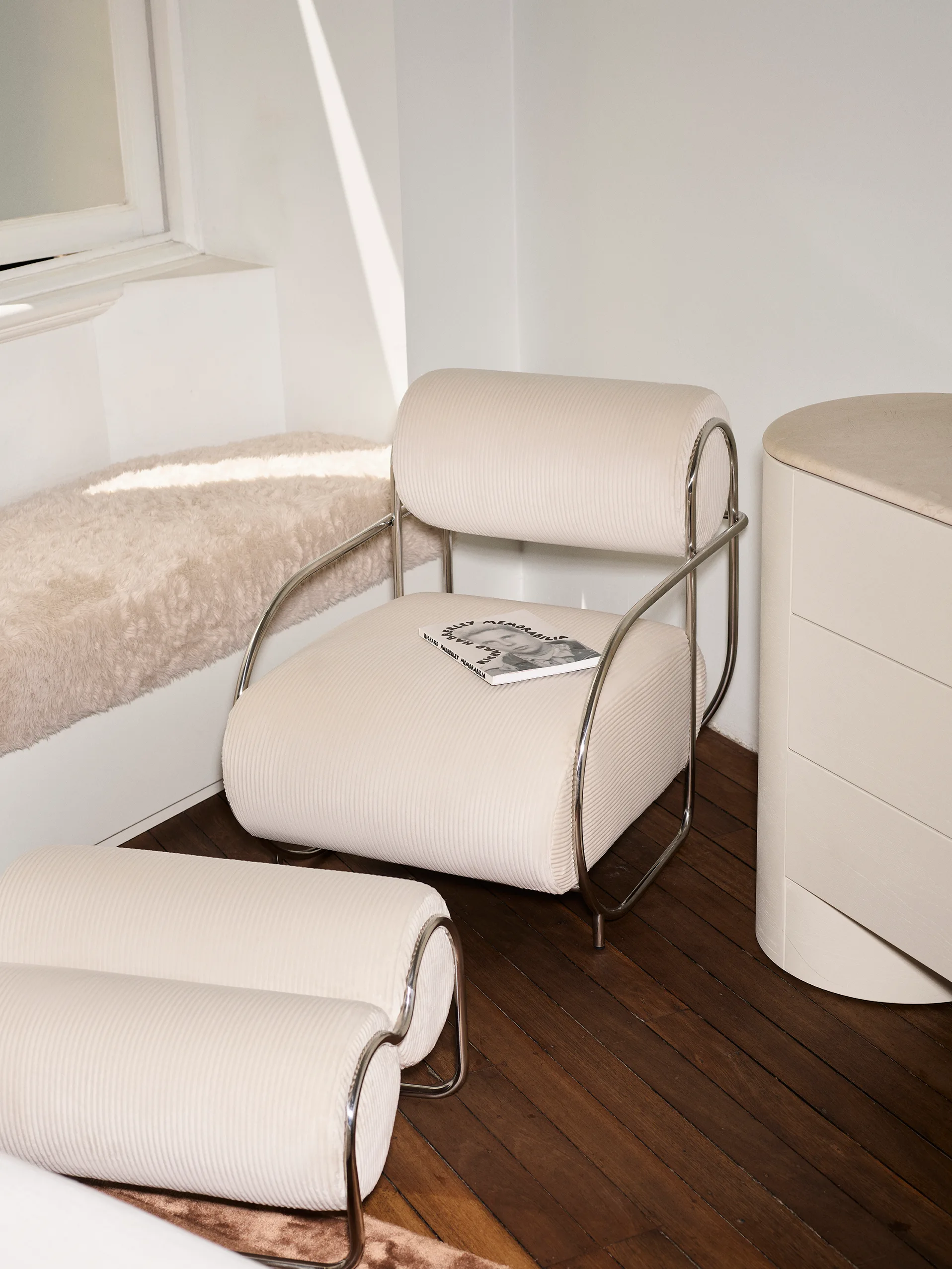 A cream corduroy armchair and matching ottoman with shiny chrome frames sit on dark wood floors next to a curved cabinet and a window ledge.