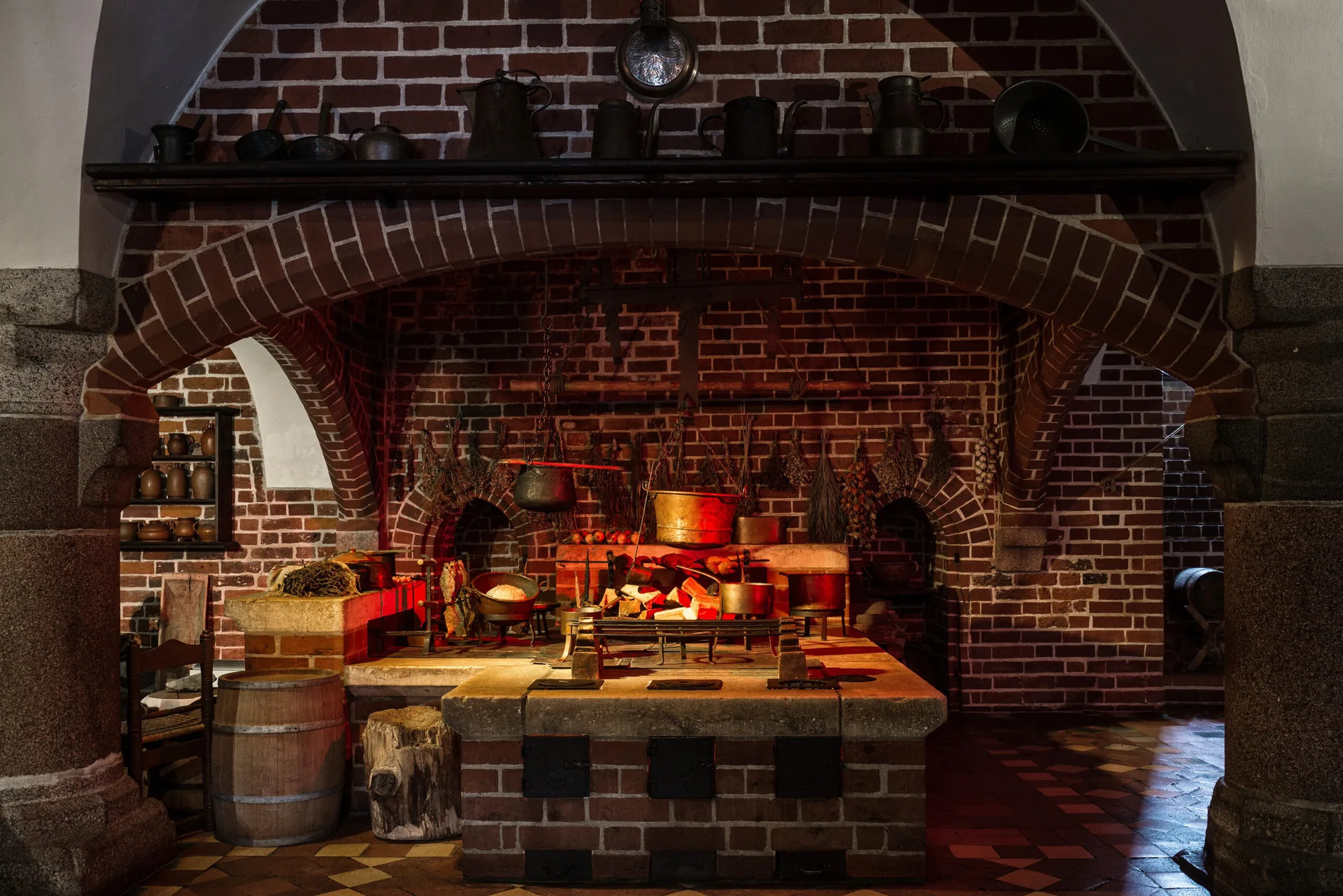 A rustic brick kitchen hearth features a thick stone counter, arched brick alcoves, hanging copper pots, and a wooden barrel on a tiled floor.