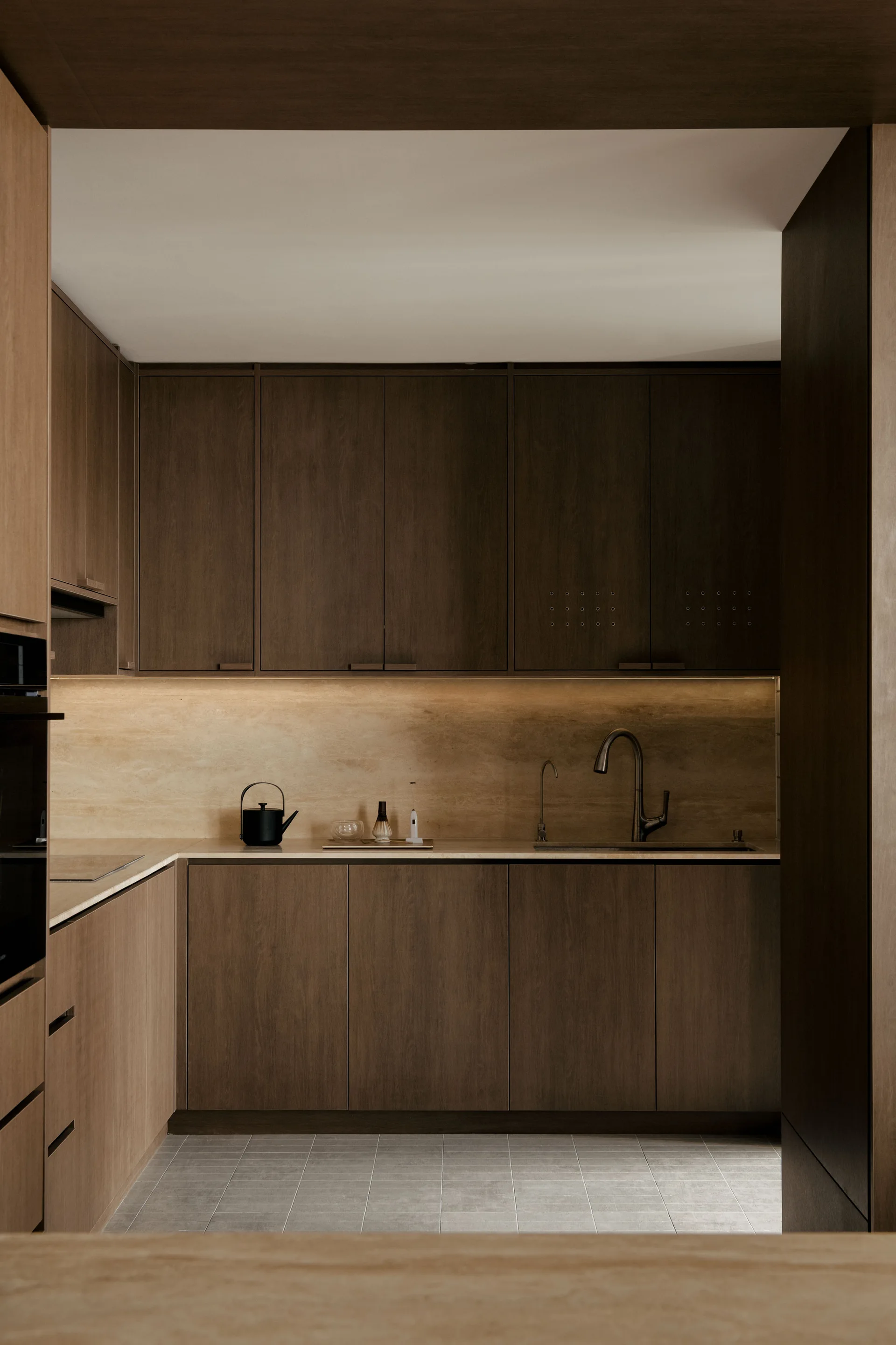 A minimalist kitchen featuring dark wood cabinetry, a light stone slab backsplash with warm under-cabinet lighting, and grey tiled flooring.