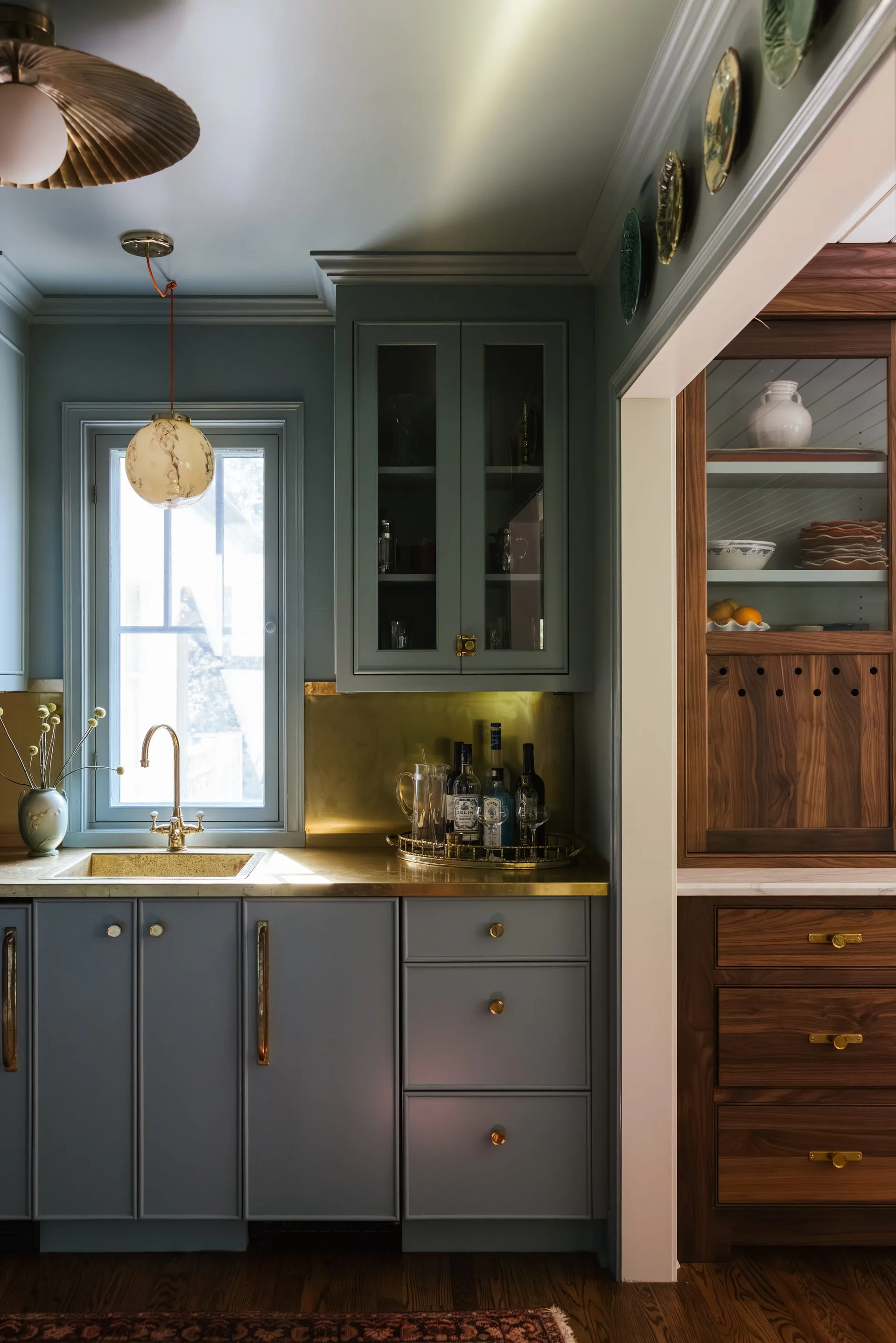 A kitchen wet bar with blue-grey cabinets, a solid brass countertop and backsplash, and an adjacent walnut wood pantry cabinet.