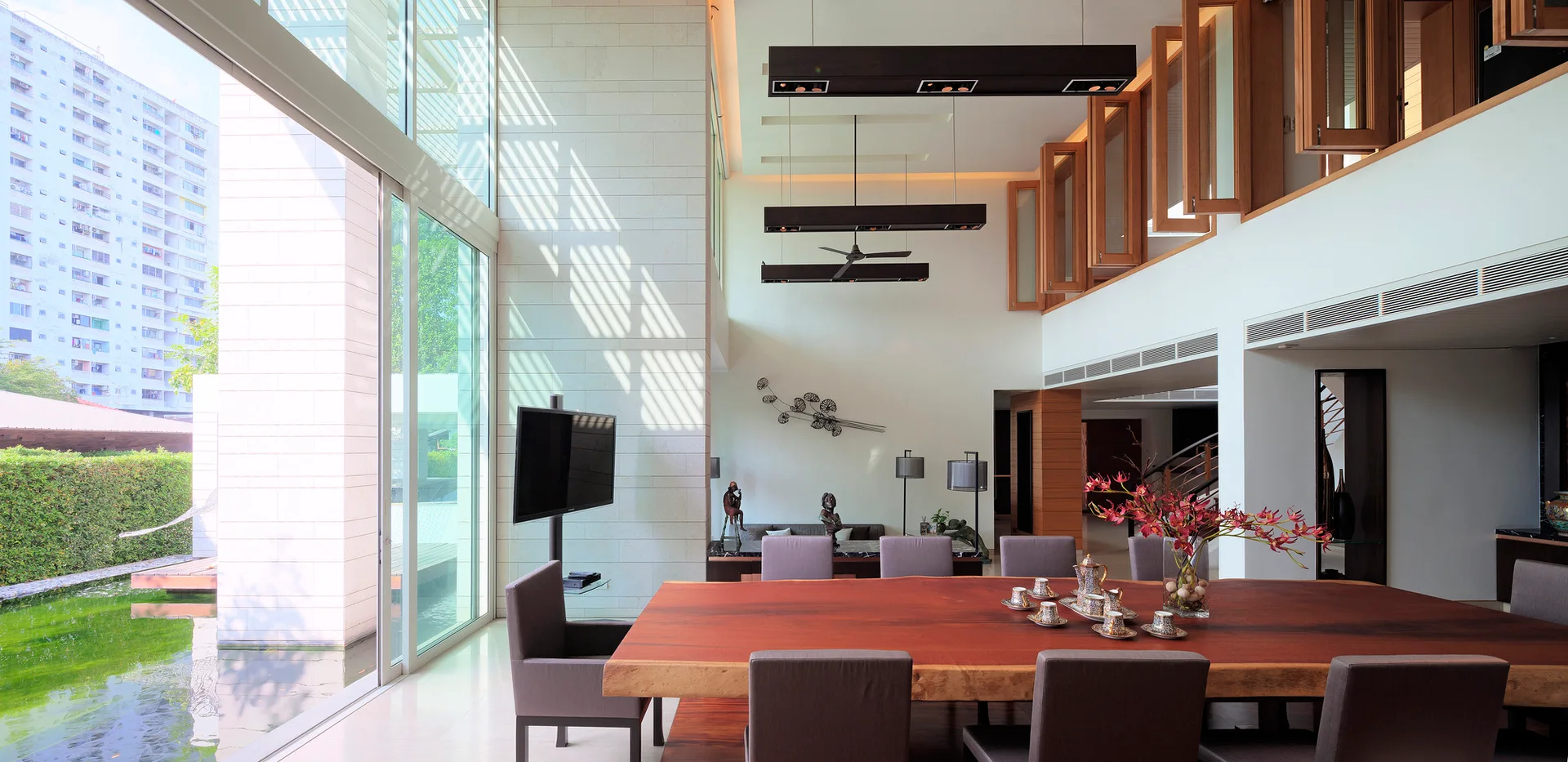A modern double-height dining room features a large live-edge wood table, grey chairs, and a glass wall overlooking an outdoor pond.