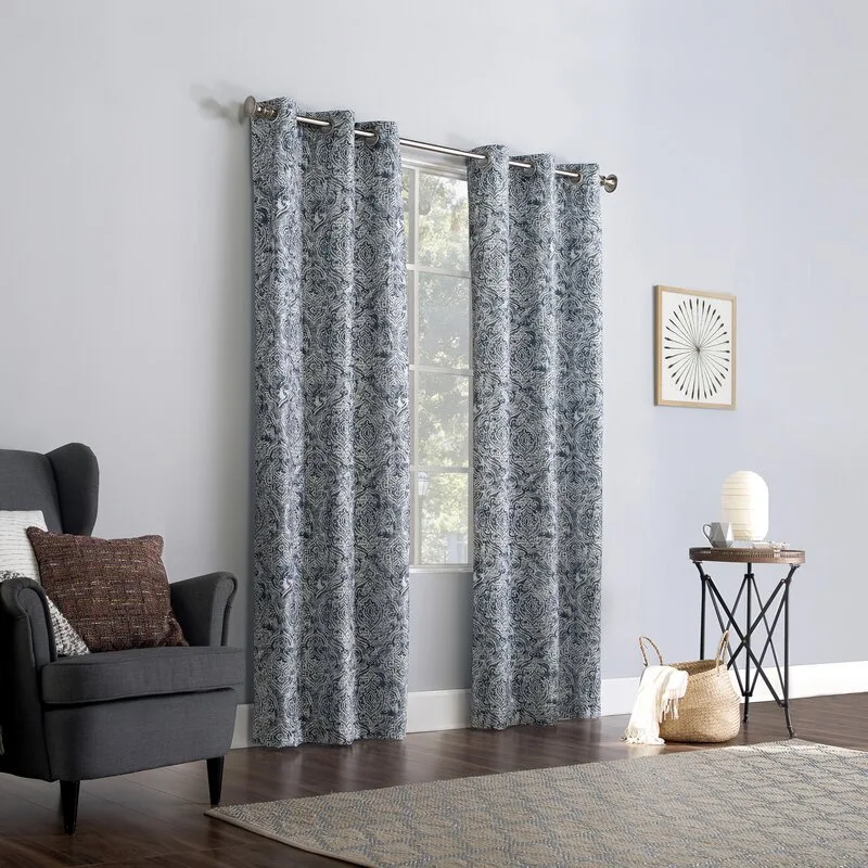 Blue and white patterned grommet curtains hang over a window next to a grey wingback armchair on dark wood floors.