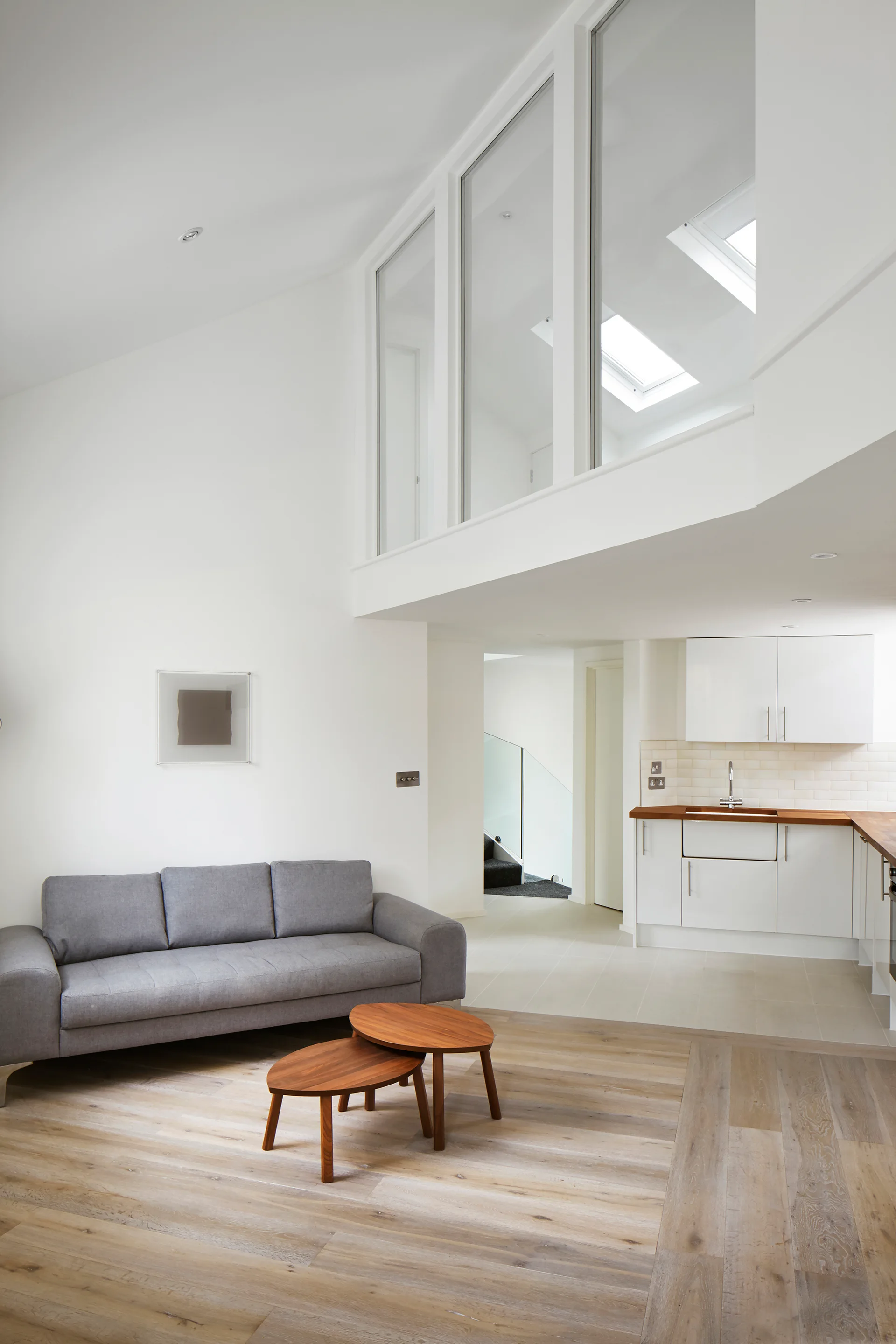 A bright open-plan living room features a grey sofa, nested wood coffee tables, and an upper loft window beneath a sloped ceiling with skylights.