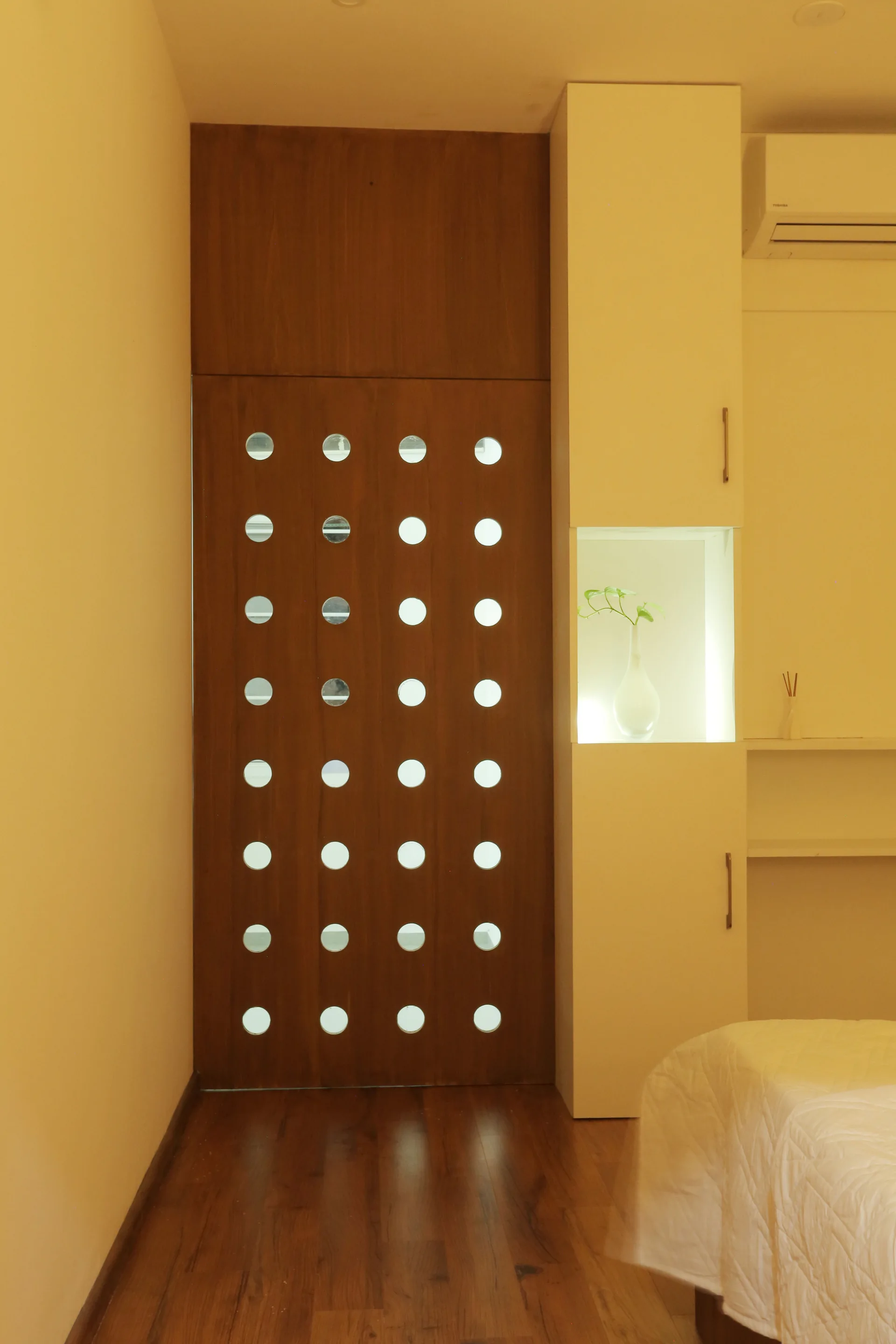A dark wood door with a grid of circular cutouts stands next to a white built-in cabinet featuring an illuminated display niche.