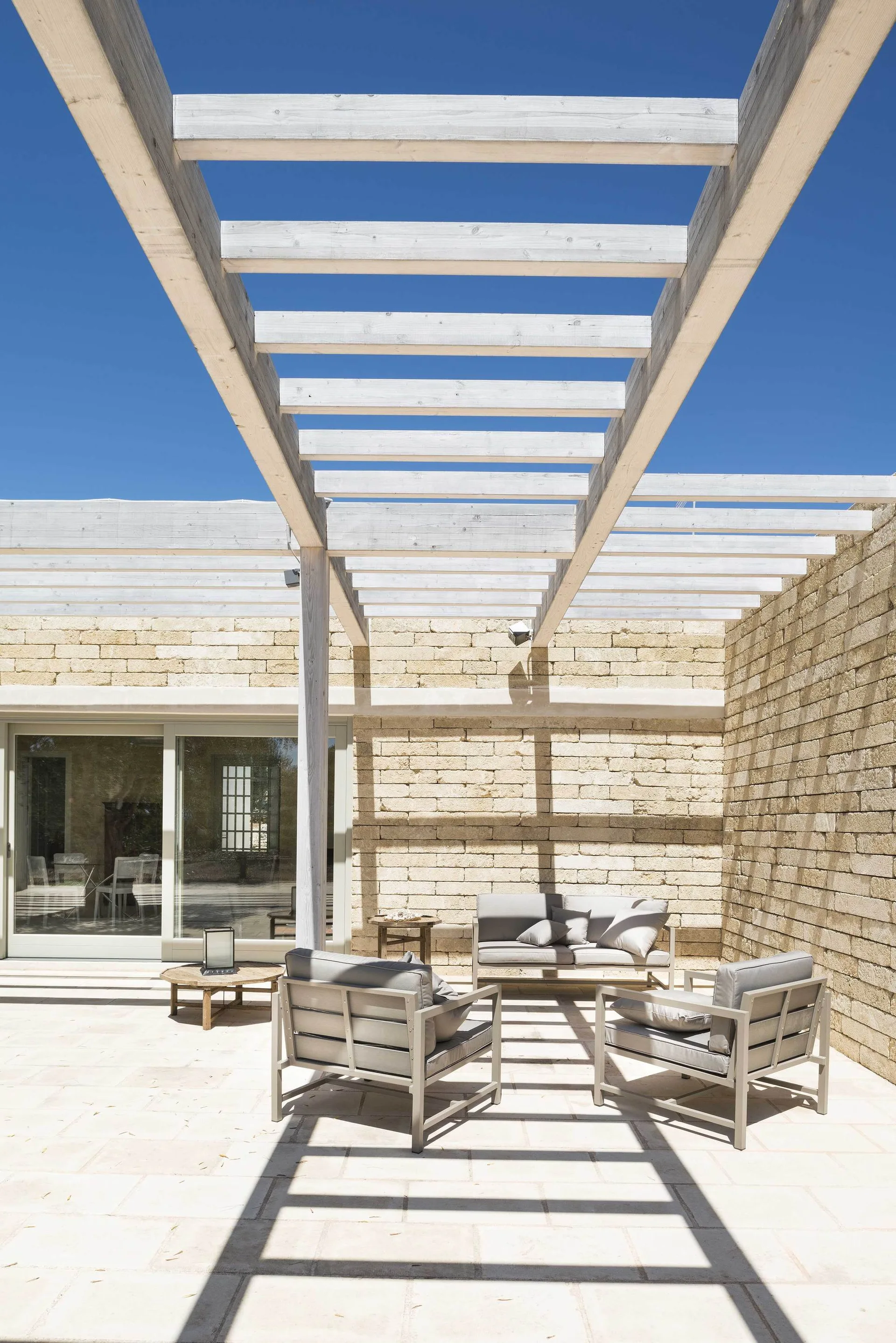 A sunny paved outdoor patio features a gray seating set arranged beneath a white wood pergola next to a textured stone block wall.