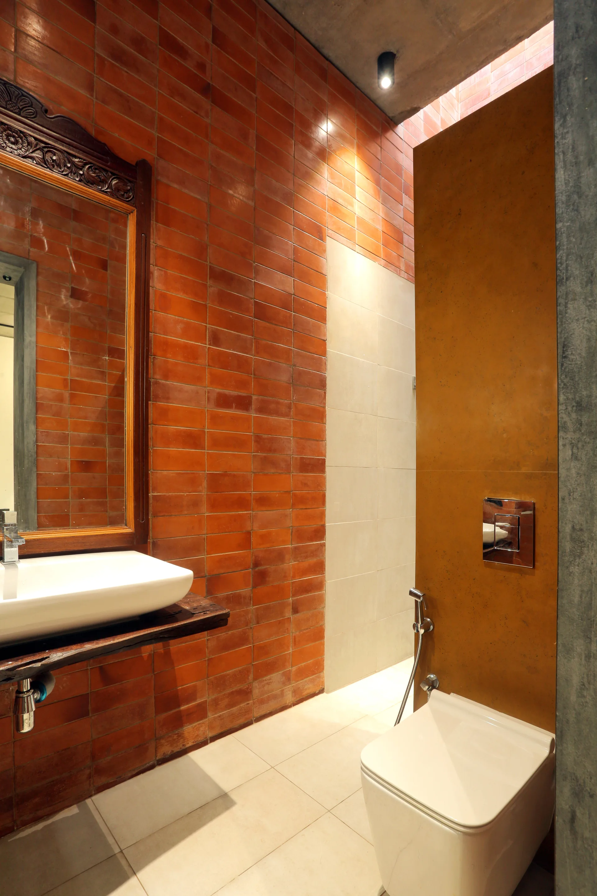 A modern bathroom features a terracotta brick wall, a white vessel sink on a raw wood slab, an ornate wood mirror, and a wall-mounted toilet.