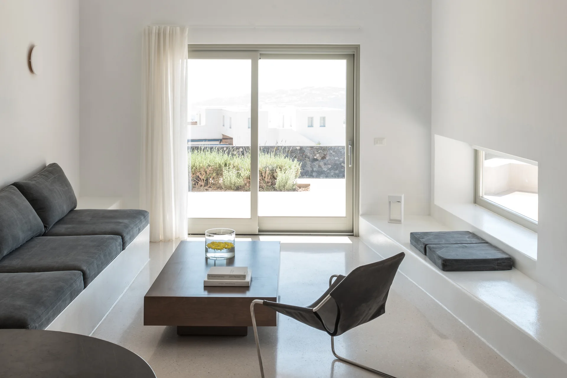 A minimalist white living room features a built-in plaster sofa with dark grey cushions, a blocky dark wood coffee table, and a large sliding glass door.