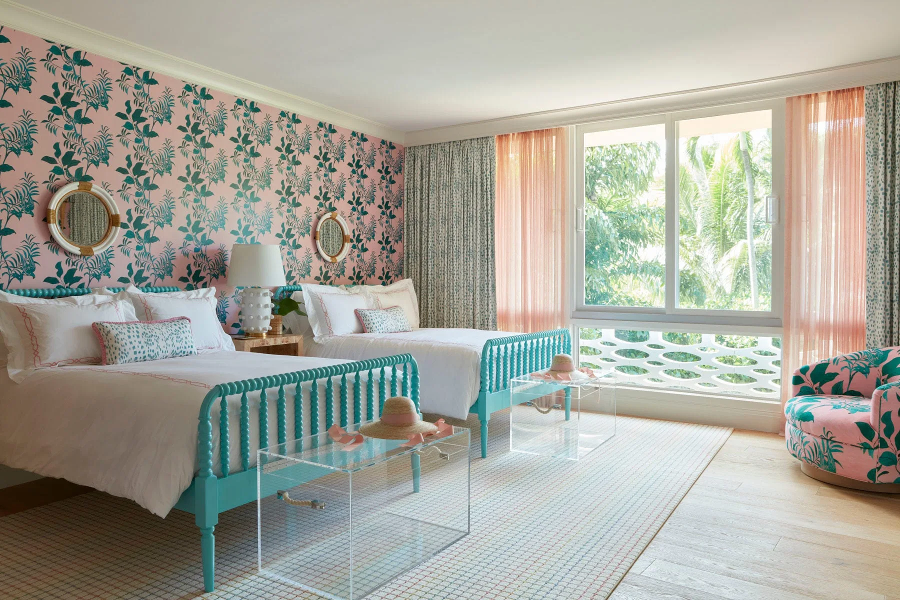 A bright bedroom features two teal spindle twin beds, pink botanical wallpaper, and clear acrylic benches on a patterned rug.