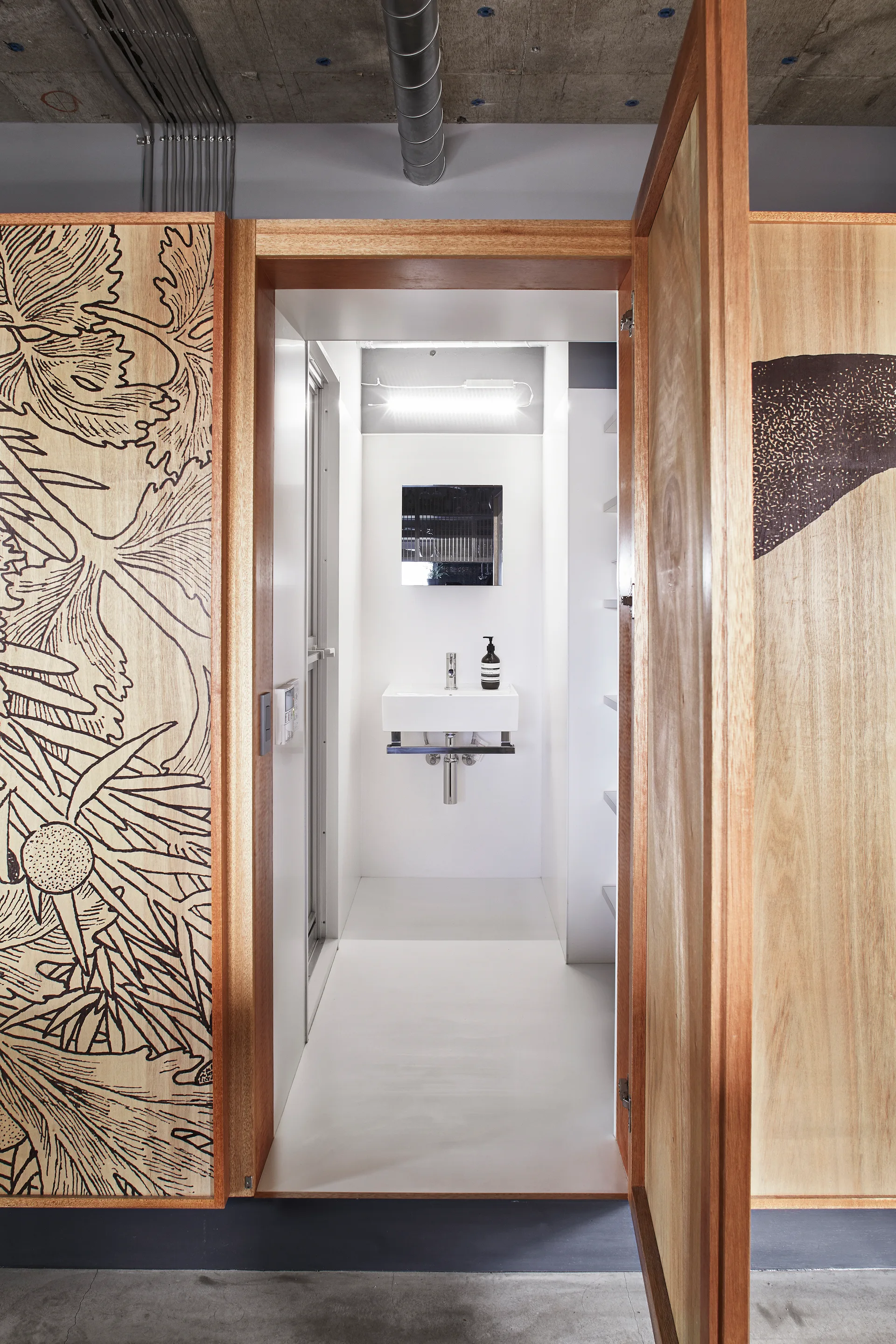 A minimalist white powder room featuring a floating sink and mirror, viewed through an open entryway with ornate engraved wood paneling.