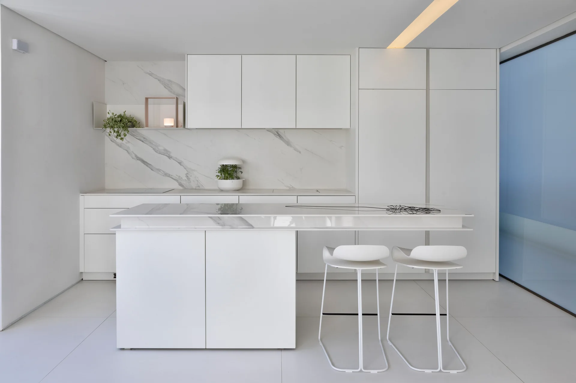 A minimalist white kitchen features a marble backsplash, seamless slab cabinets, and a white island with two contemporary bar stools.