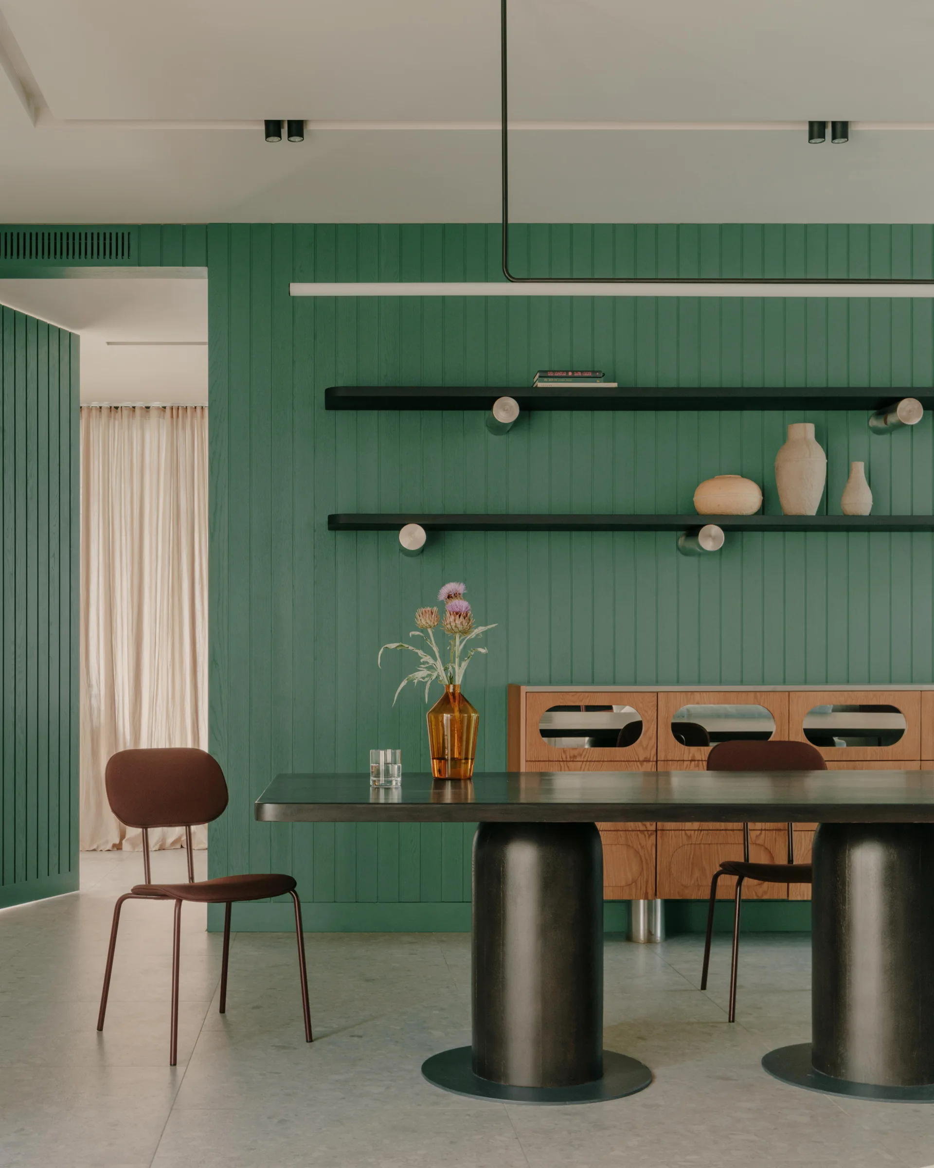 A dining room featuring a black table with cylindrical legs, burgundy chairs, and floating shelves against a green vertical-planked wood wall.