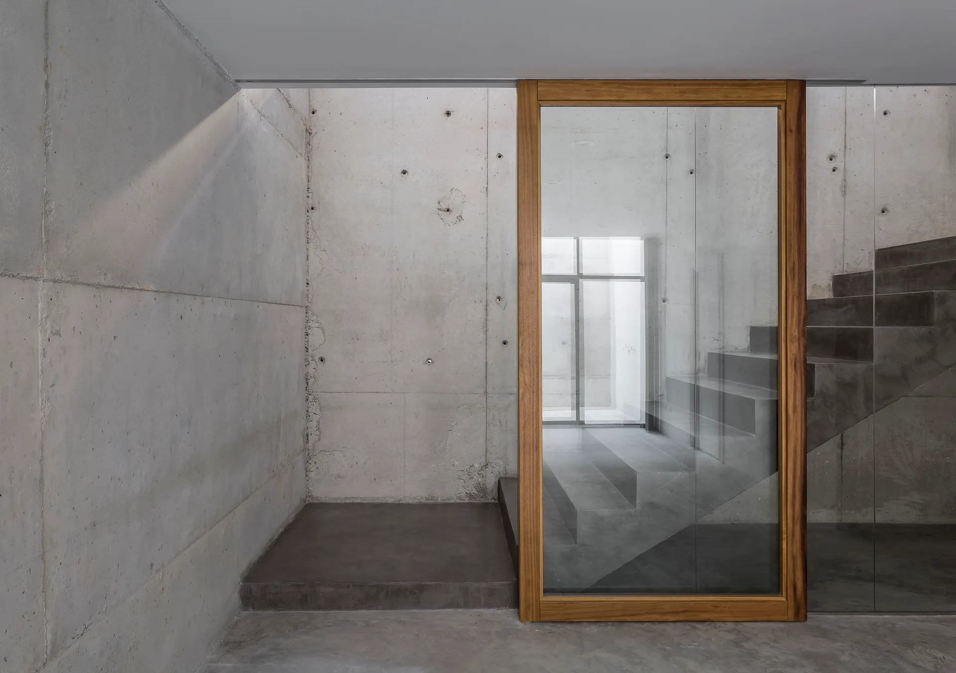 Exposed concrete walls line a modern stairwell with a large wood-framed glass door opening to grey concrete stairs.