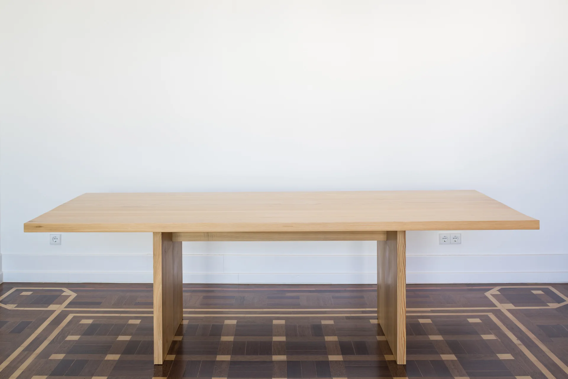 A minimalist light wood dining table with solid panel legs stands on an intricate dark wood geometric parquet floor.