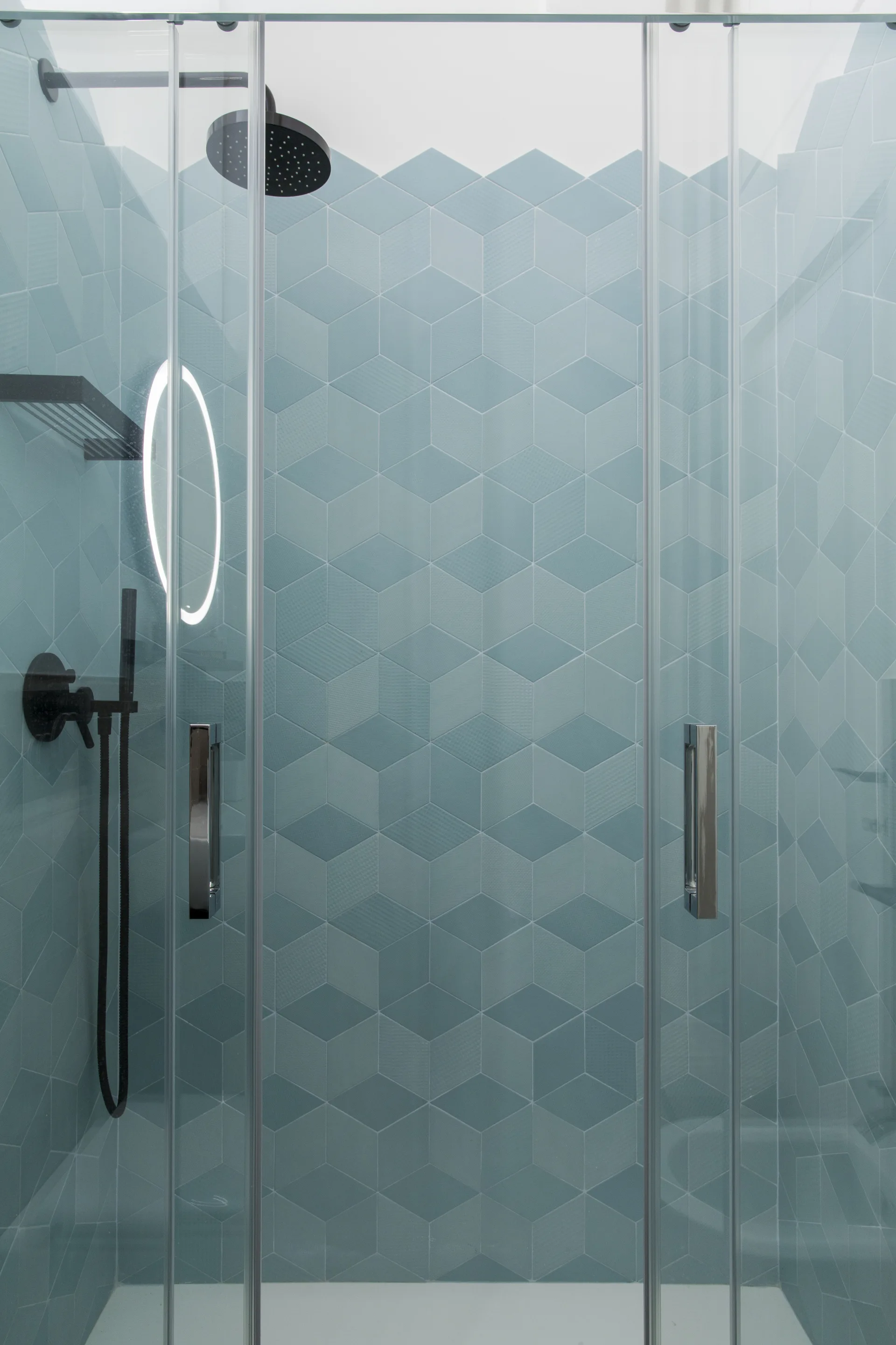 A modern walk-in shower features teal geometric cube-patterned tiles on the wall, matte black fixtures, and sliding glass doors.