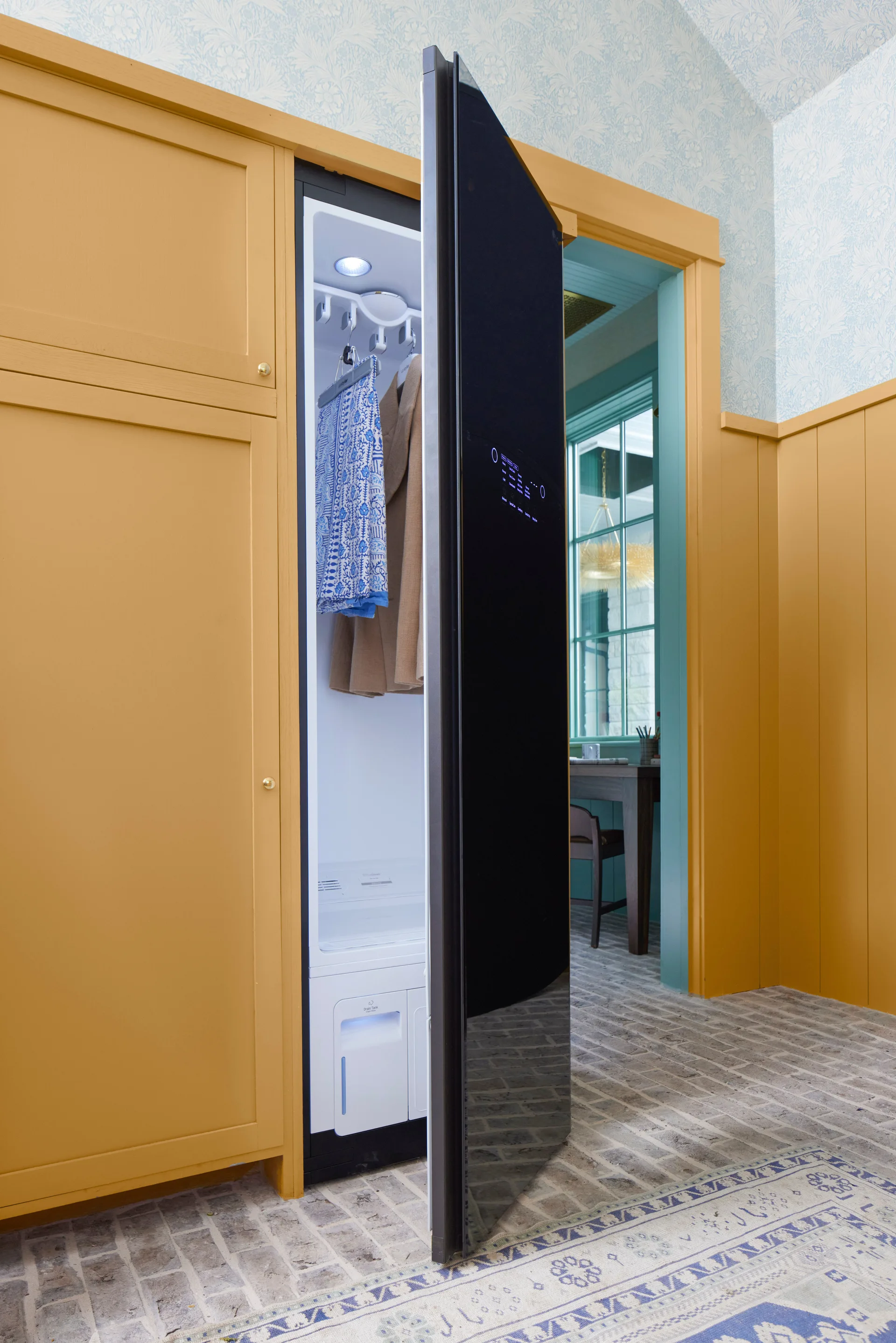 A tall black clothing care appliance stands open inside mustard yellow built-in cabinetry on a herringbone brick floor next to blue floral wallpaper.