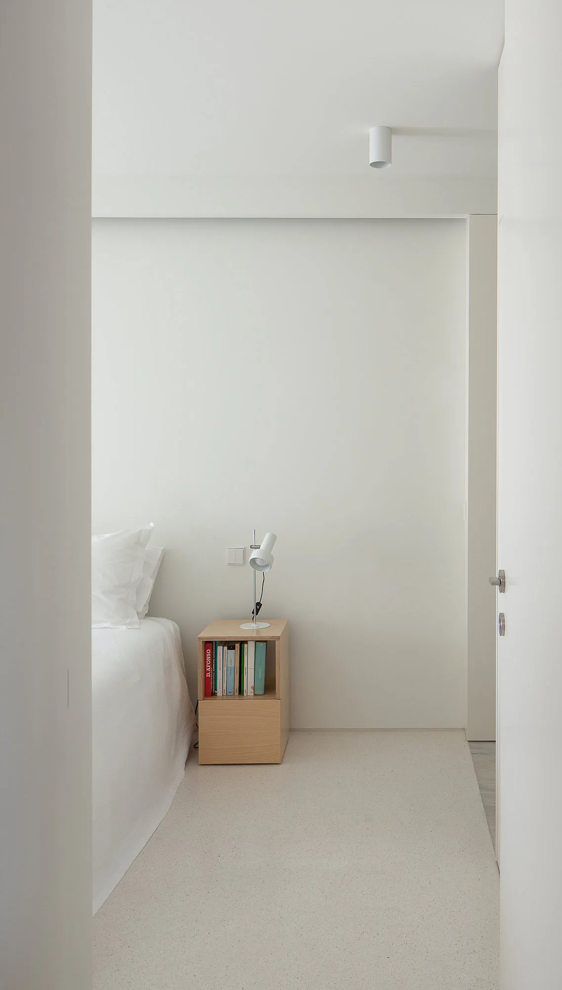 A minimalist white bedroom featuring a white bed, a light wood cube nightstand with books, and a slim white desk lamp.