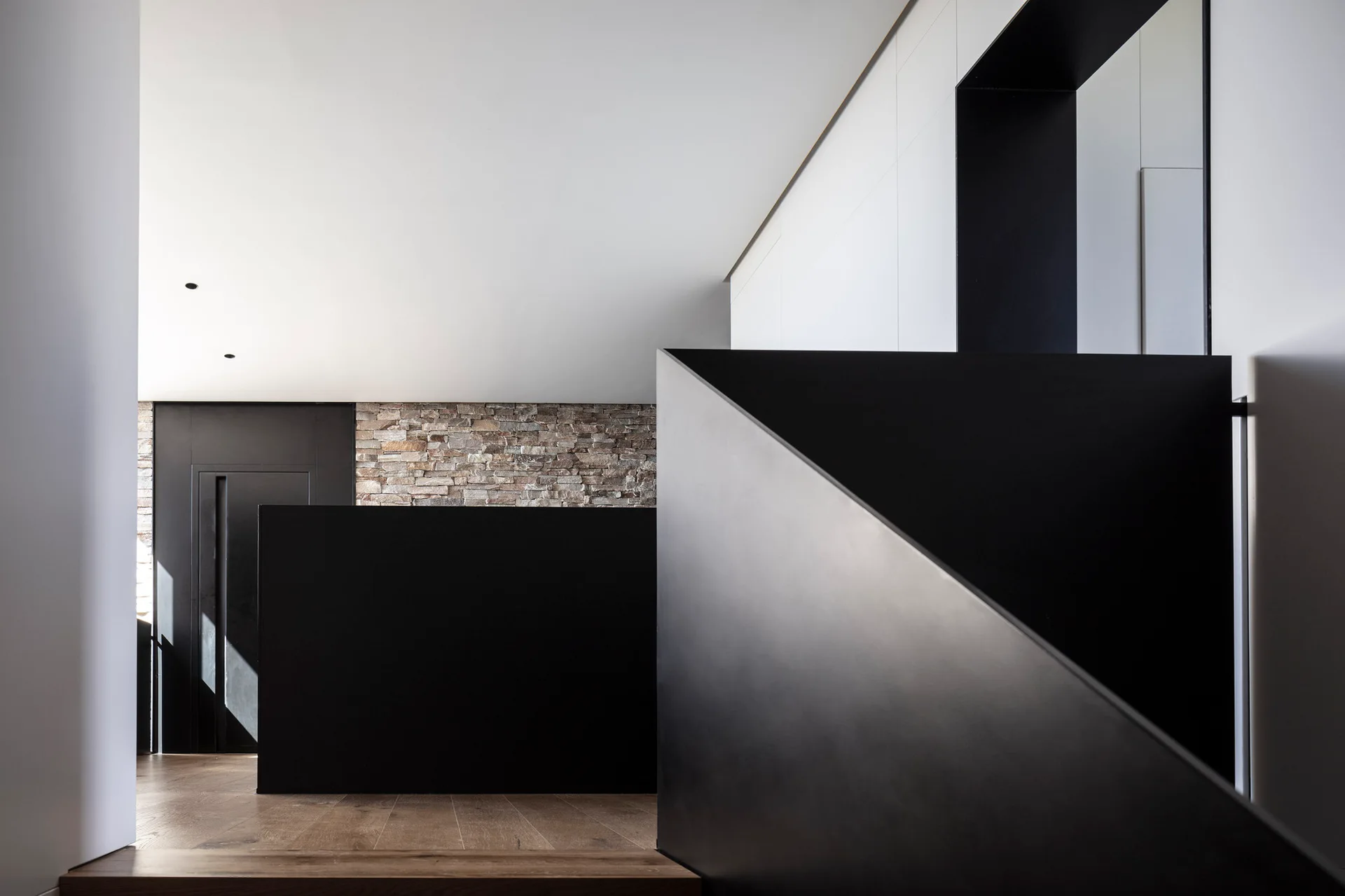 A minimalist staircase landing featuring a sharp, angular black metal railing, light wood floors, and a textured natural stone wall.