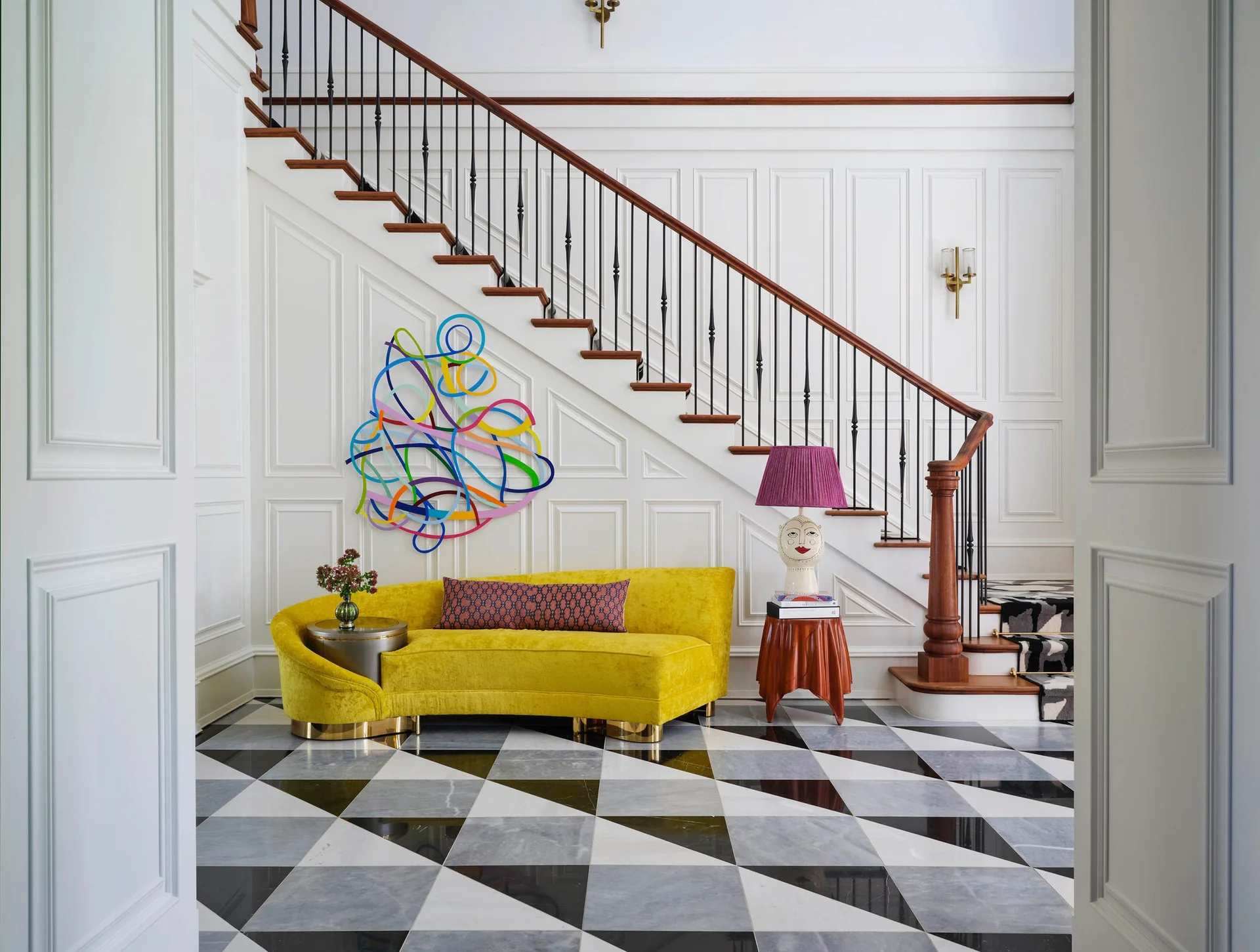 An entryway foyer features a bright yellow curved velvet sofa, a staircase with black metal railing, and a black, white, and grey geometric marble floor.