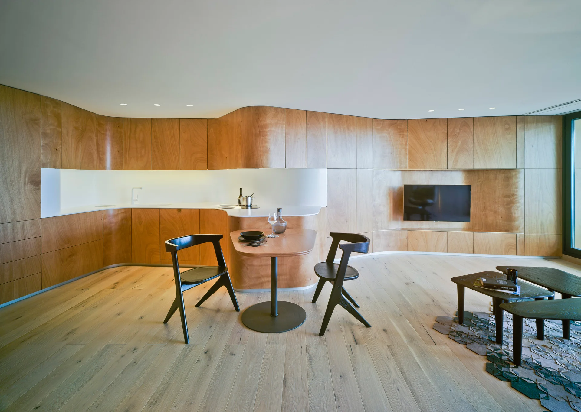 An open concept apartment features a curved wood paneled wall integrating kitchen cabinets and a flat-screen television above light wood floors.