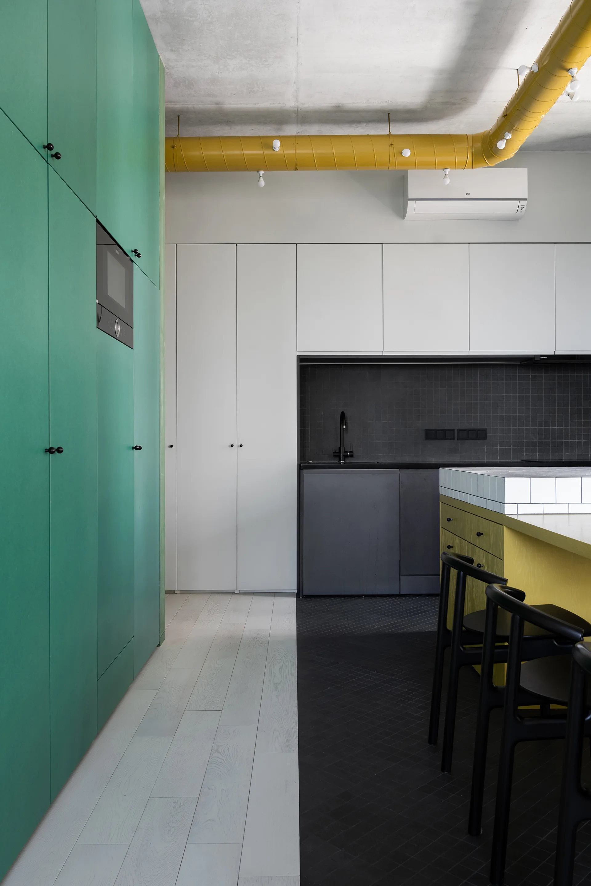 A modern kitchen features green and white floor-to-ceiling cabinets, a yellow island, and a split wood and black tile floor.