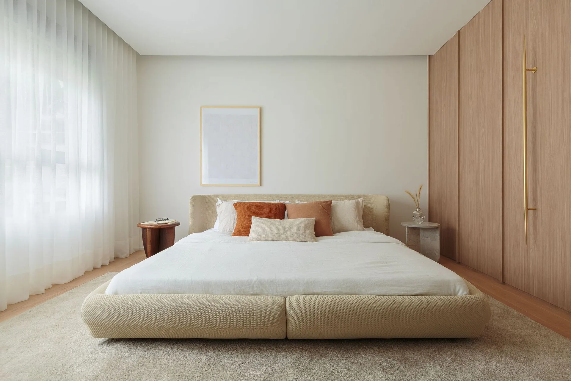 A minimalist bedroom features a low-profile upholstered bed flanked by mismatched side tables, sheer white curtains, and a wood-paneled wall.