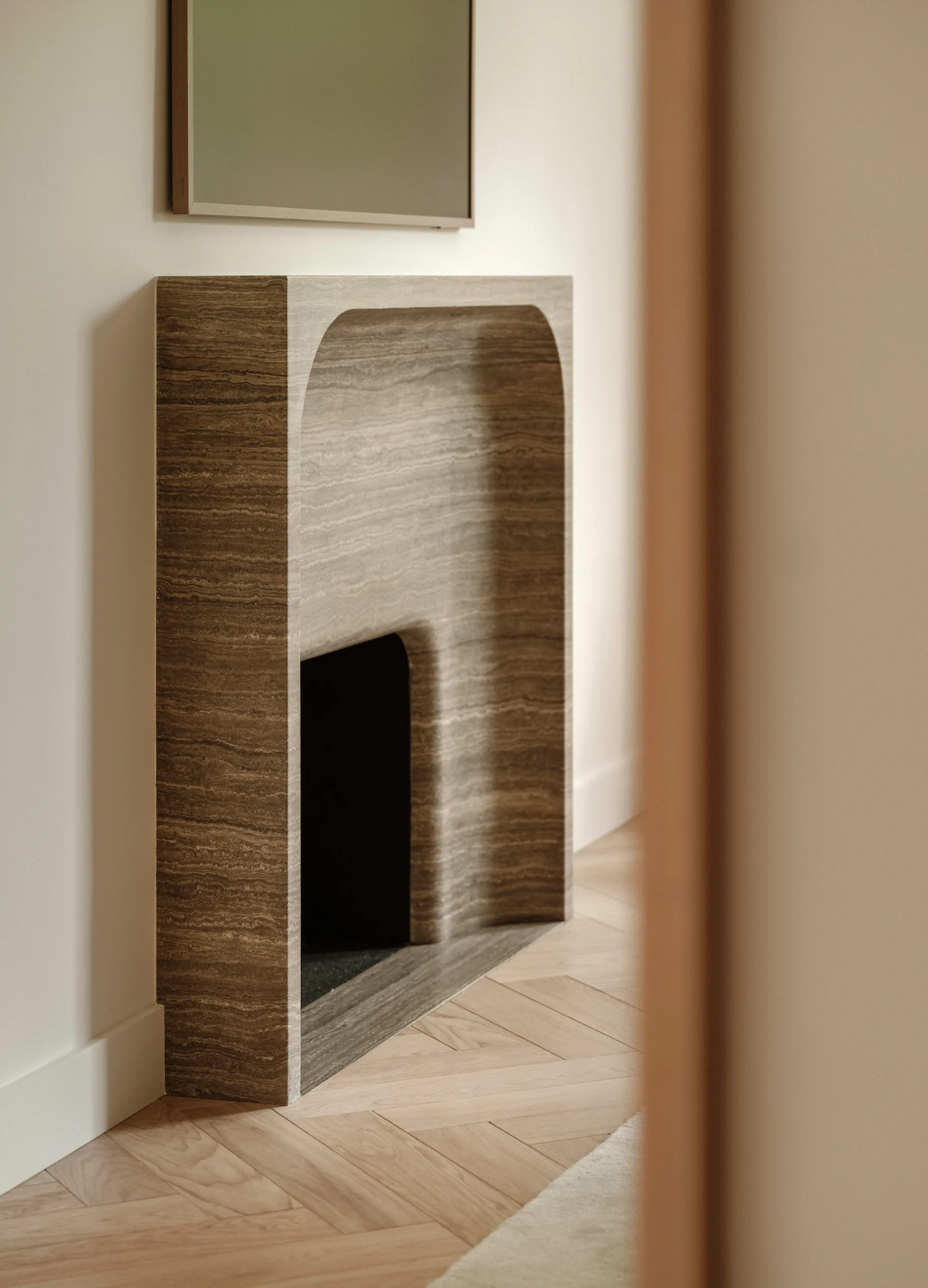 A minimalist brown veined stone fireplace surround with an arched opening sits on a light wood herringbone patterned floor against an off-white wall.