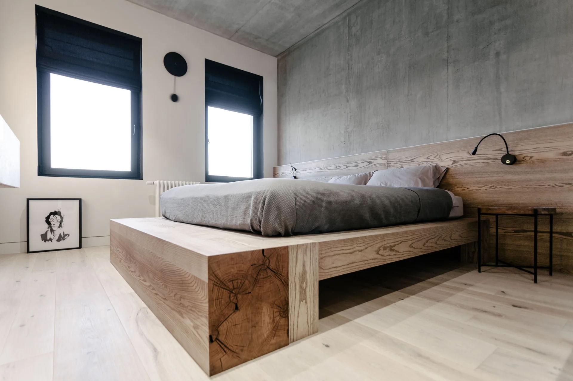 A low-profile light wood platform bed sits against a textured grey concrete wall in a minimalist bedroom with pale wood floors.