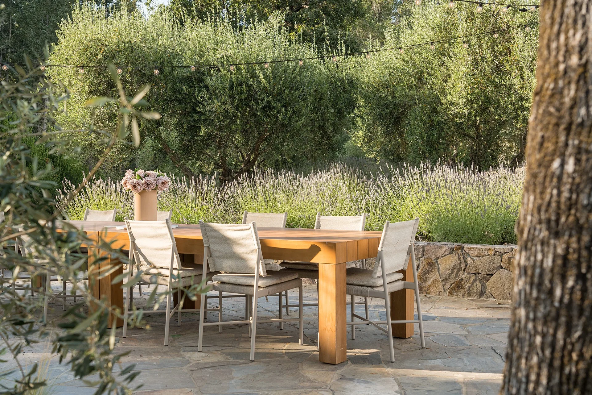 A large wooden dining table and beige chairs sit on an irregular flagstone patio next to a stone wall and blooming lavender.