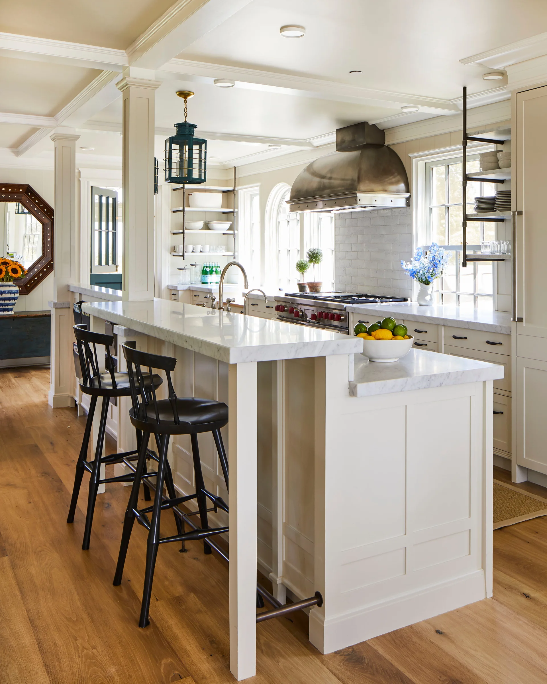 A bright kitchen features a white marble-topped island with two black bar stools, cream cabinets, a metal range hood, and light hardwood floors.