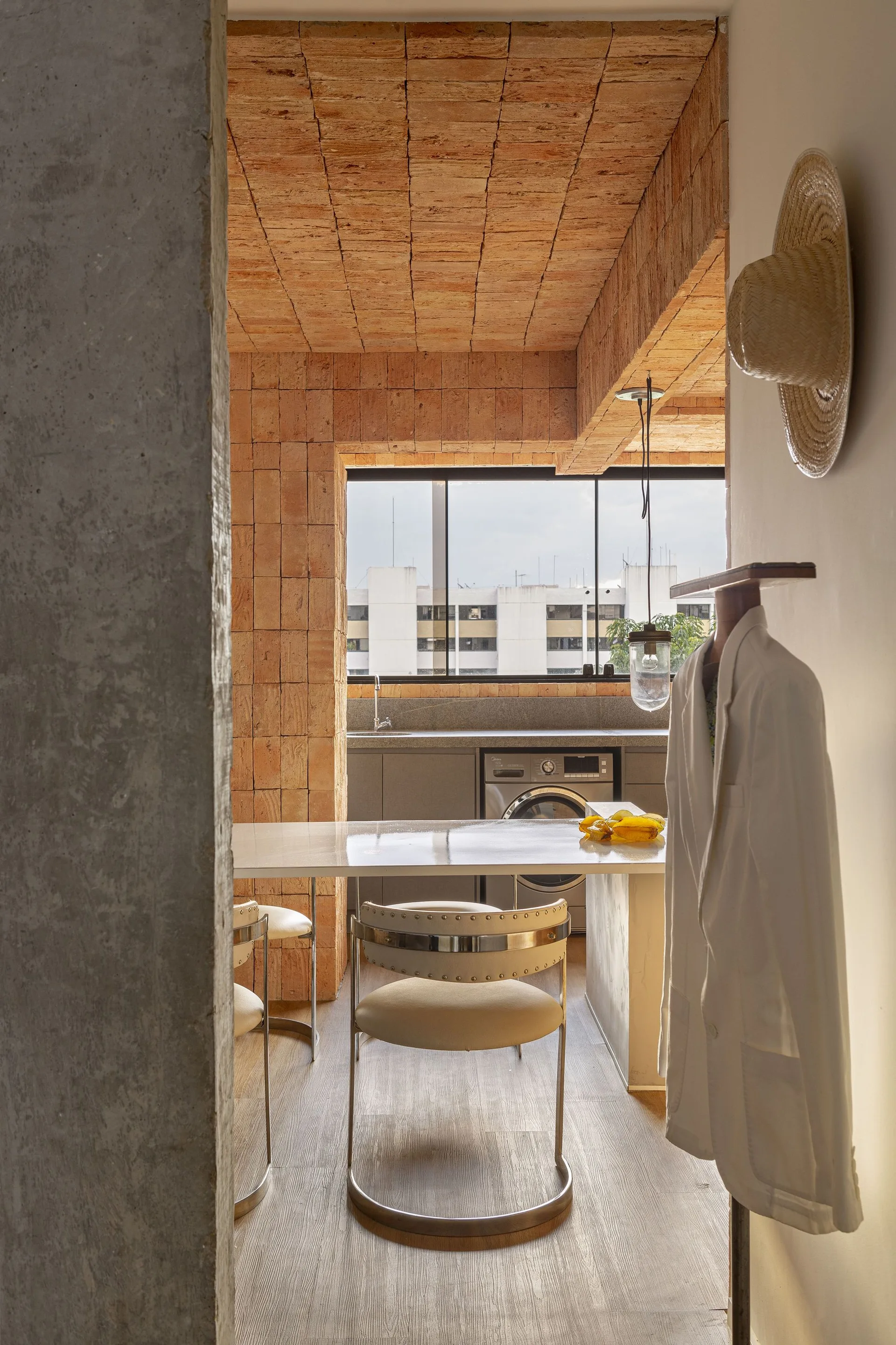 A modern kitchen and dining area features an exposed terracotta brick ceiling, a concrete pillar, a white dining table with metal chairs, and grey cabinets.