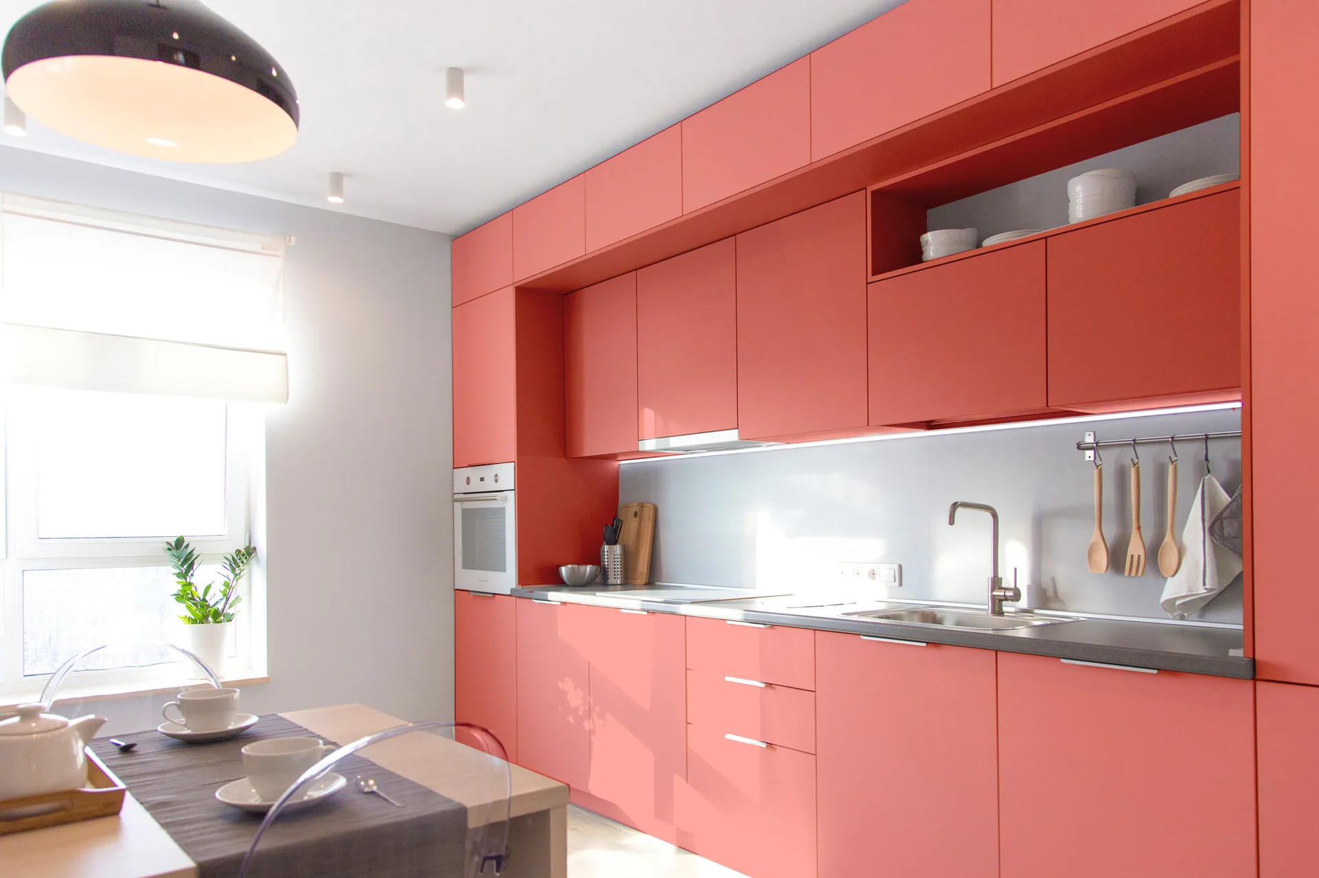 A modern kitchen with matte salmon pink handleless cabinets, a grey backsplash, and a dining table in the foreground.