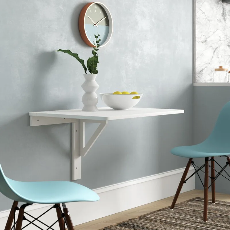 A white wall-mounted folding desk is paired with two blue molded plastic chairs in a light blue minimalist room.