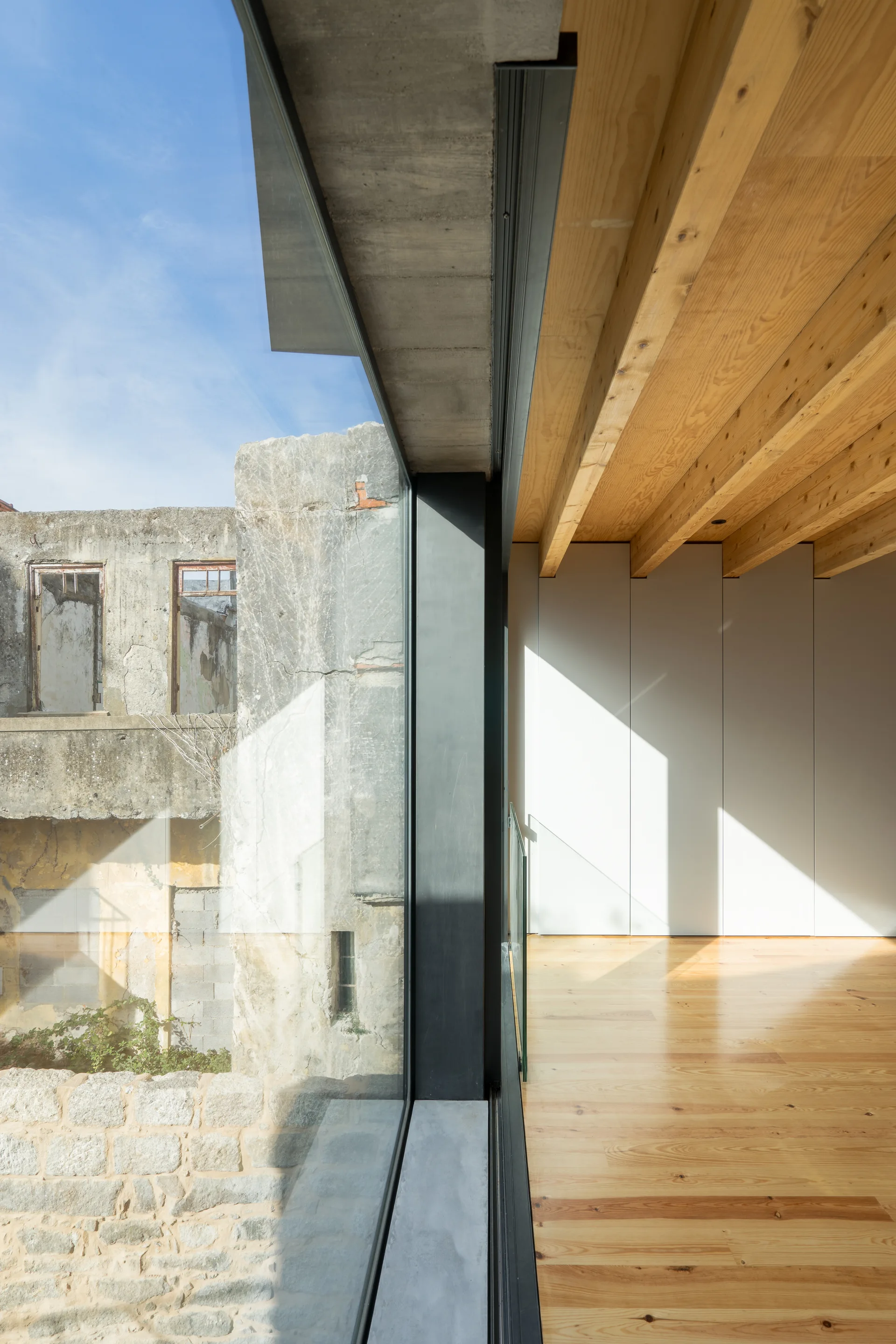 Modern interior featuring light wood flooring, exposed timber ceiling beams, white flush cabinetry, and a large window overlooking an old stone structure.