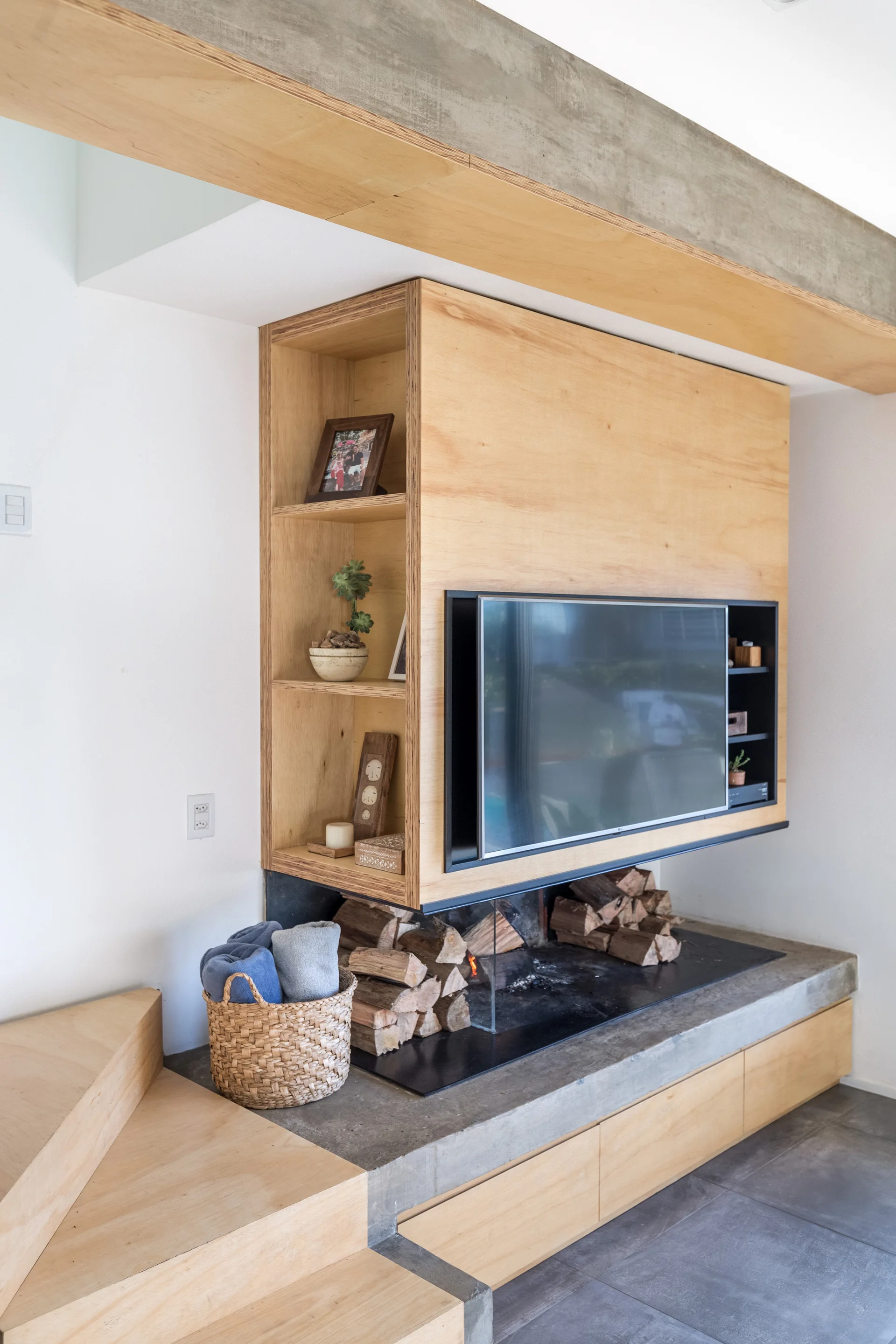 A custom plywood cabinet with a flat-screen television and open shelves is suspended over a concrete fireplace hearth stacked with logs.