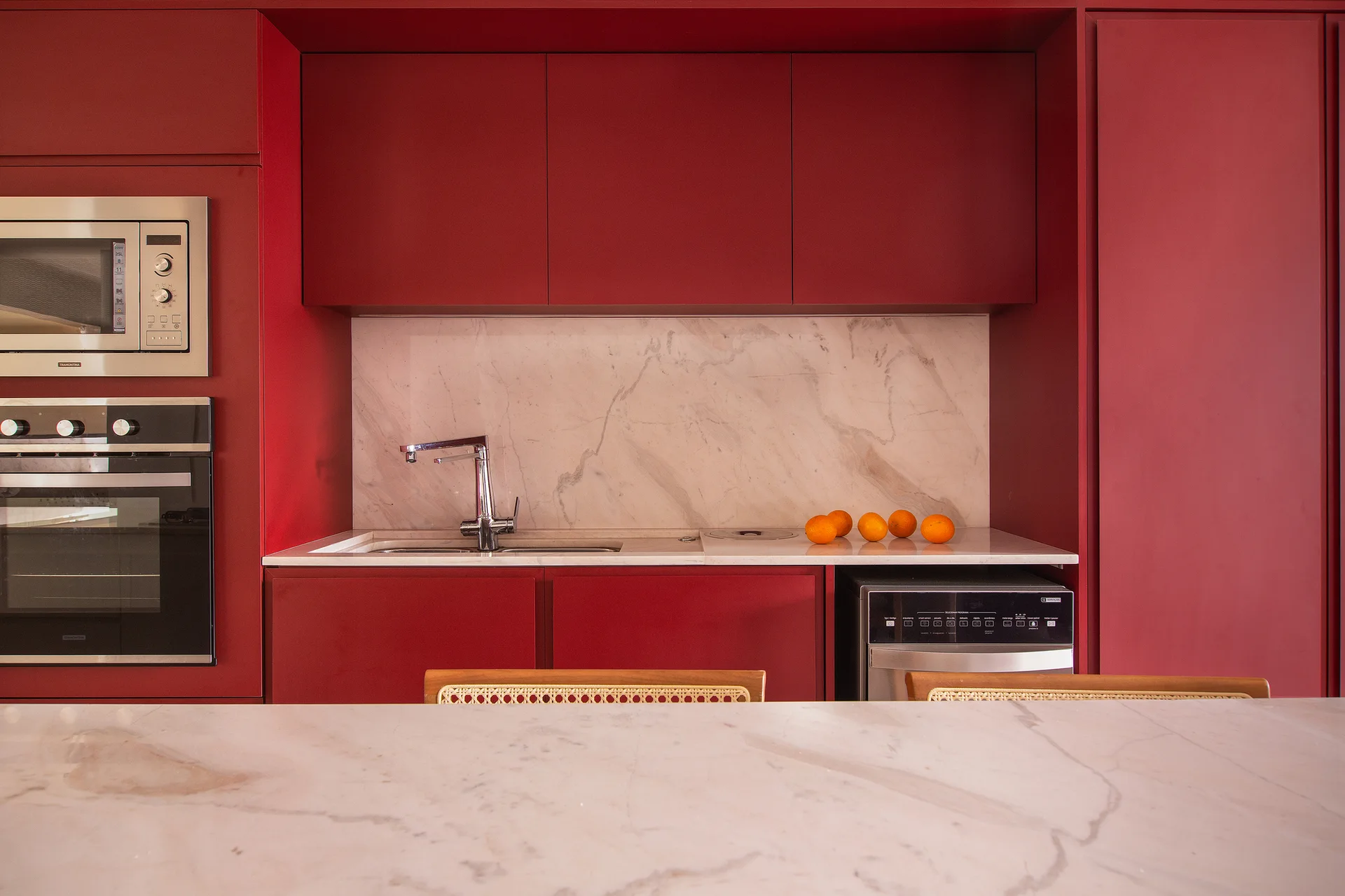 A kitchen features matte red cabinetry, a veined white marble backsplash and countertop, chrome faucet, and integrated appliances behind a marble counter.