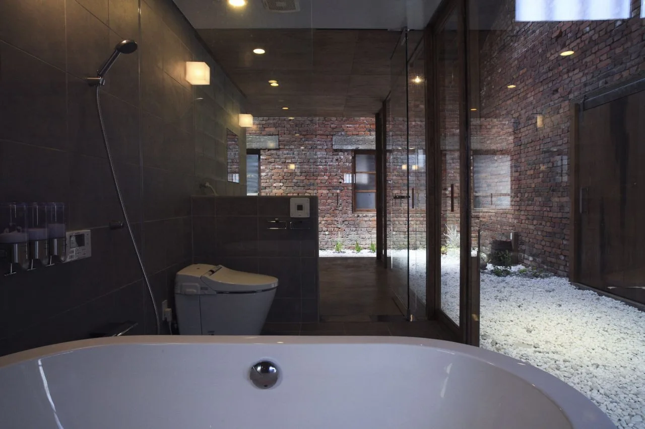 A modern bathroom with dark tiled walls, a white bathtub, and a glass wall looking out onto a brick courtyard with white gravel.
