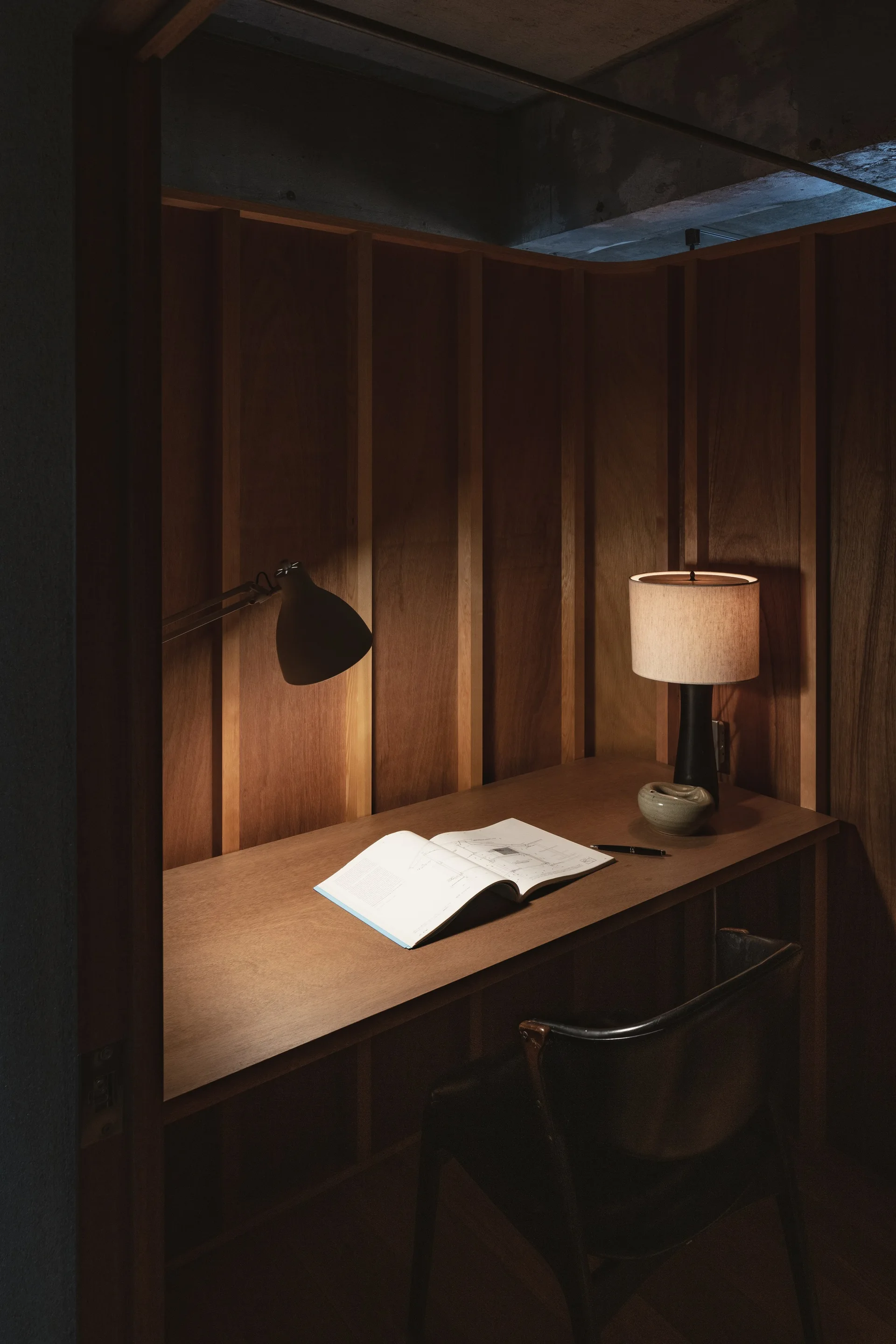 A dark wood-paneled study nook features a built-in wooden desk, a black leather chair, a table lamp, and an open book.
