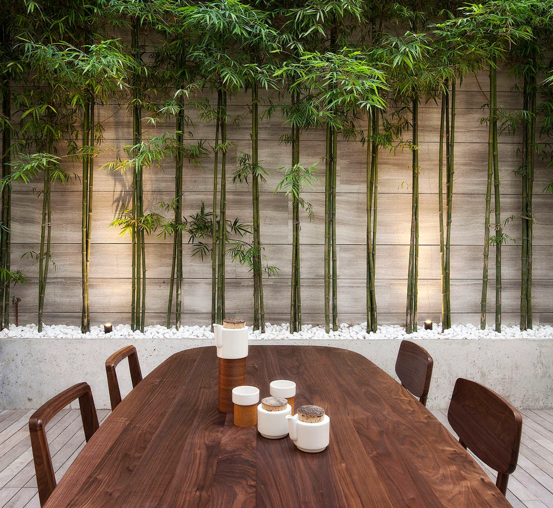A dark wood oval dining table sits on a wooden deck in front of a concrete planter filled with tall green bamboo and white gravel.