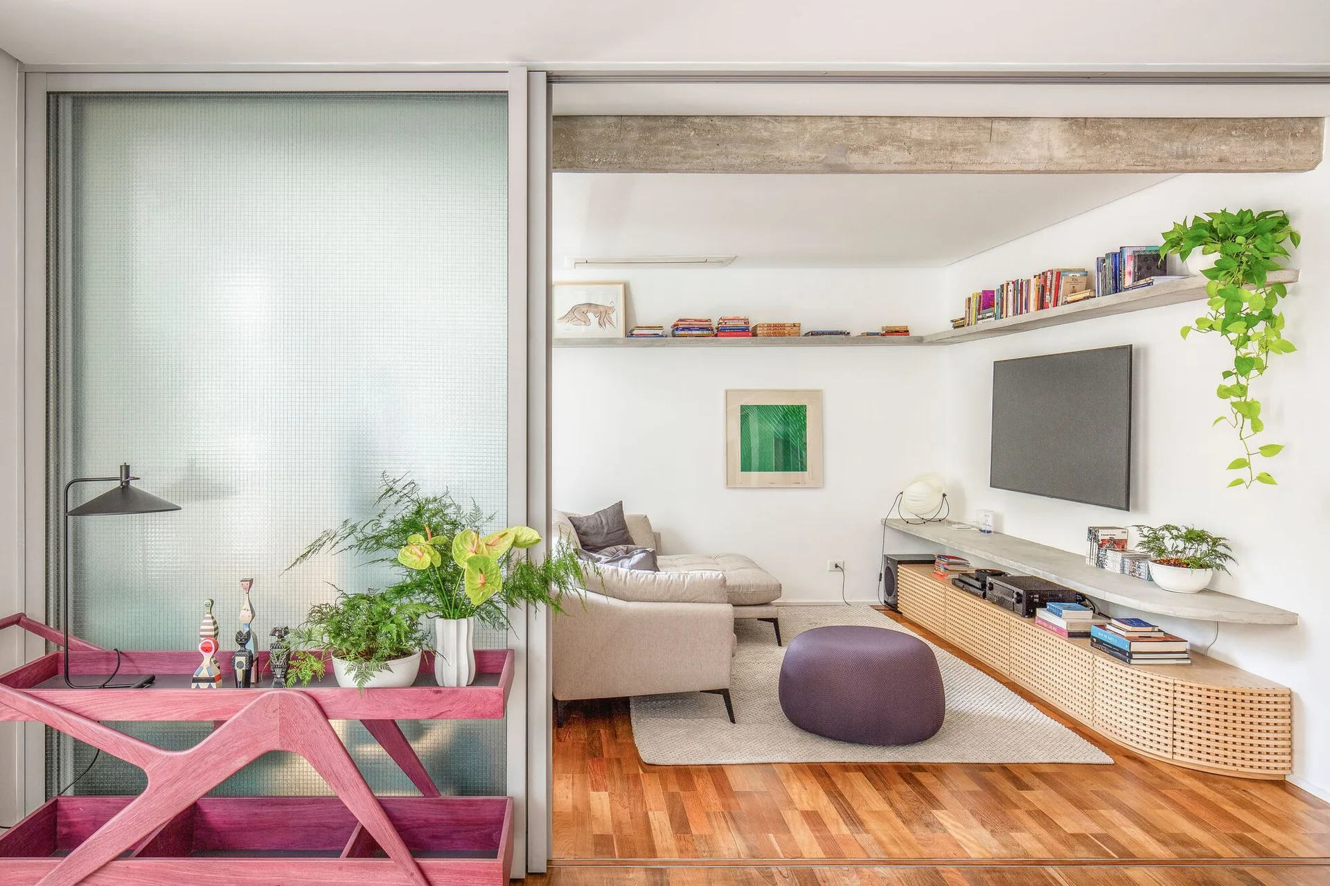 A modern living room featuring a beige sofa, a low wooden media console with woven caning, a purple ottoman, and an exposed concrete ceiling beam.