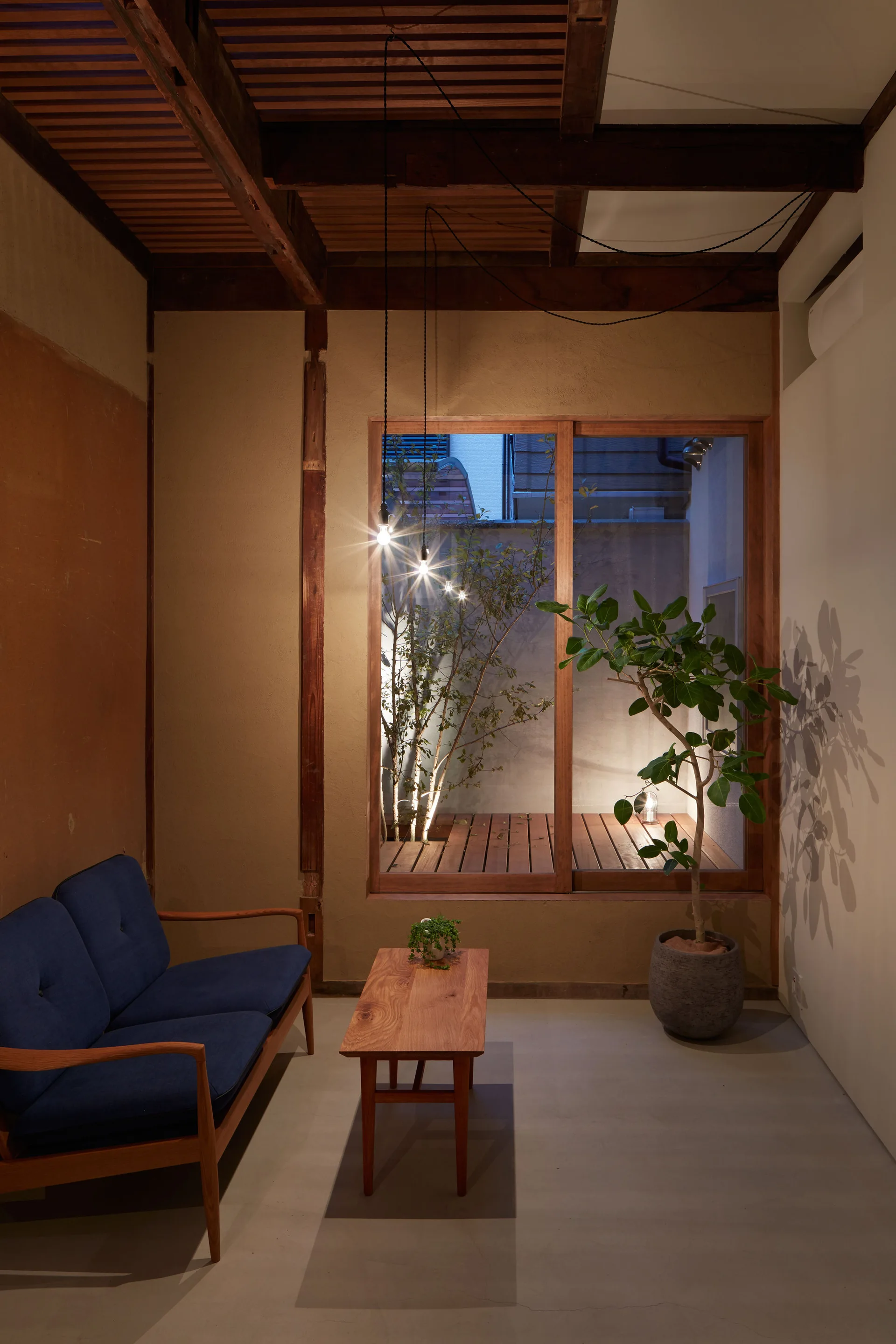A minimalist living room features a blue sofa, a wooden coffee table, and a large wood-framed window looking out onto a small lit courtyard.