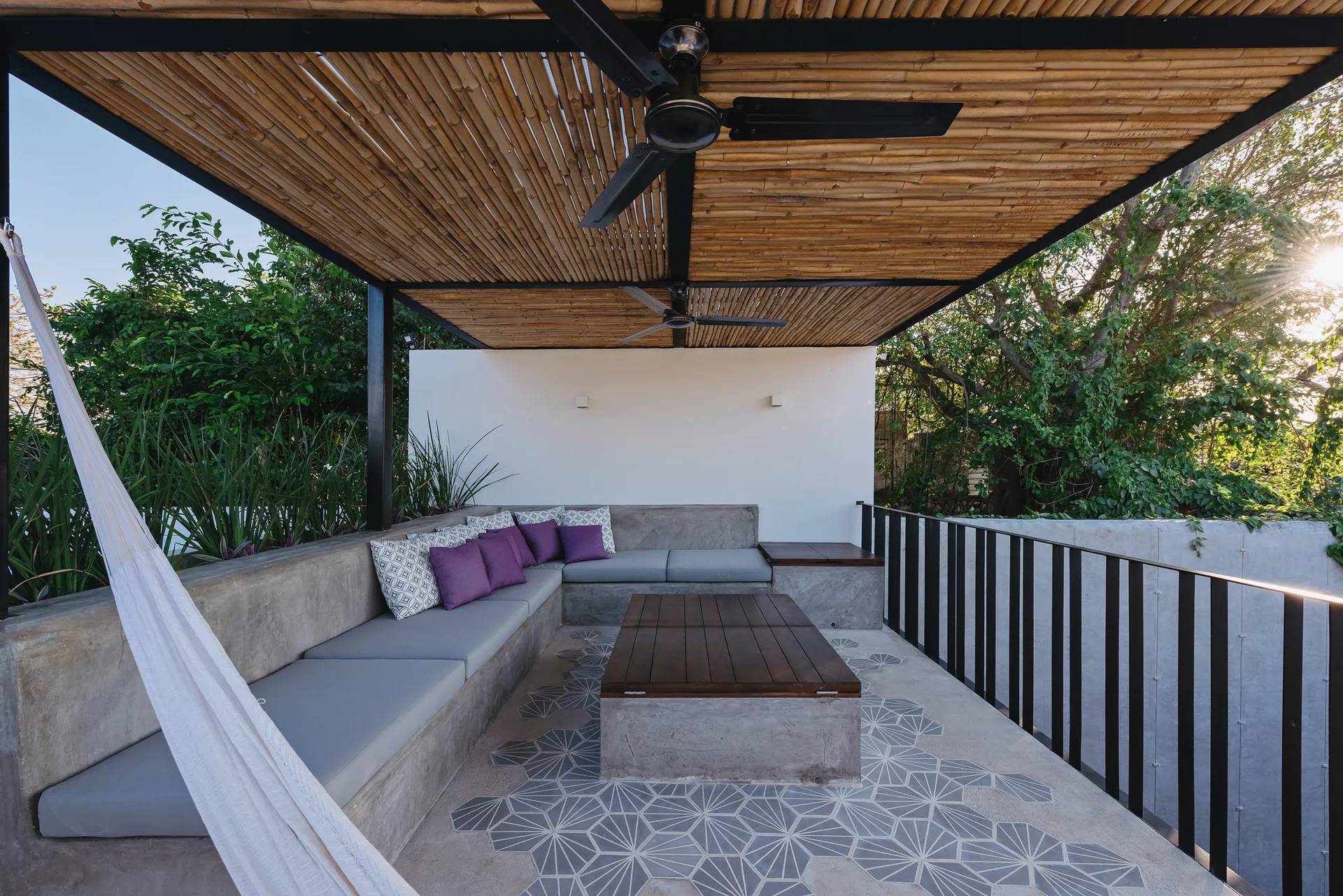 An outdoor patio features an L-shaped concrete bench with grey cushions, a bamboo slatted ceiling, and geometric tiled flooring.
