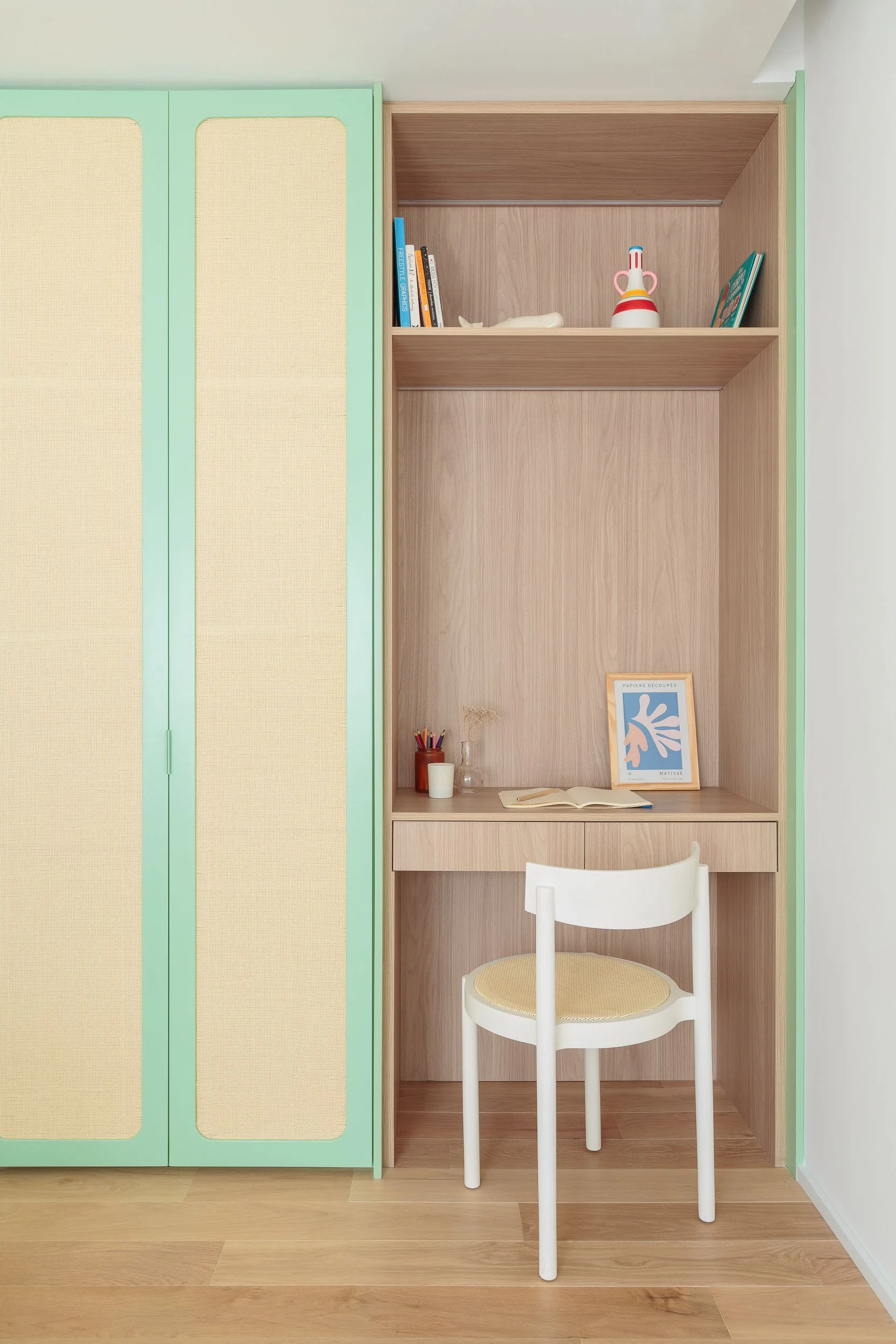 A built-in wooden desk nook with shelving next to a full-height mint green cabinet featuring woven cane panels and a white chair.