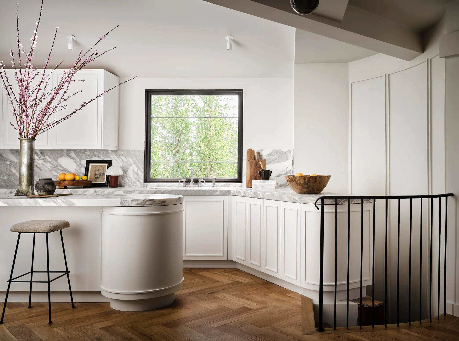 A bright white kitchen featuring shaker cabinets, a curved marble-topped island, herringbone wood floors, and a black-framed window.