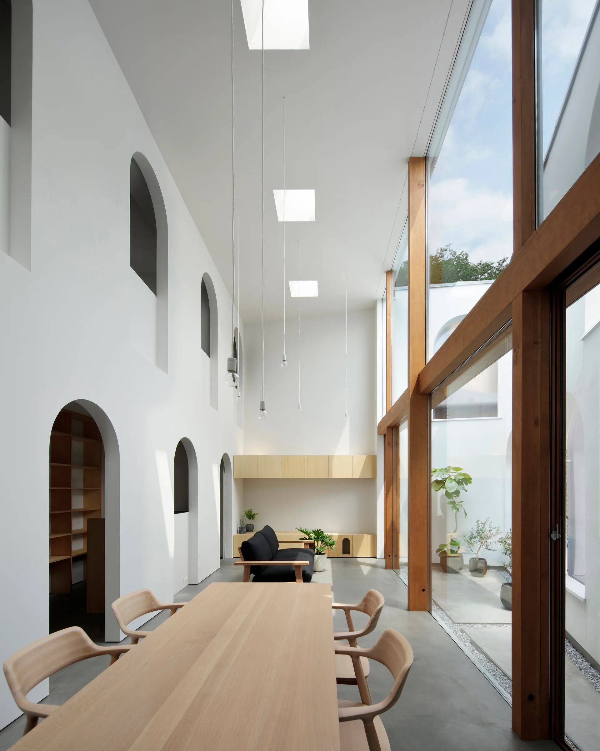 A minimalist dining and living space features a long wood table, towering arched openings, and a large wood-framed glass wall facing a courtyard.