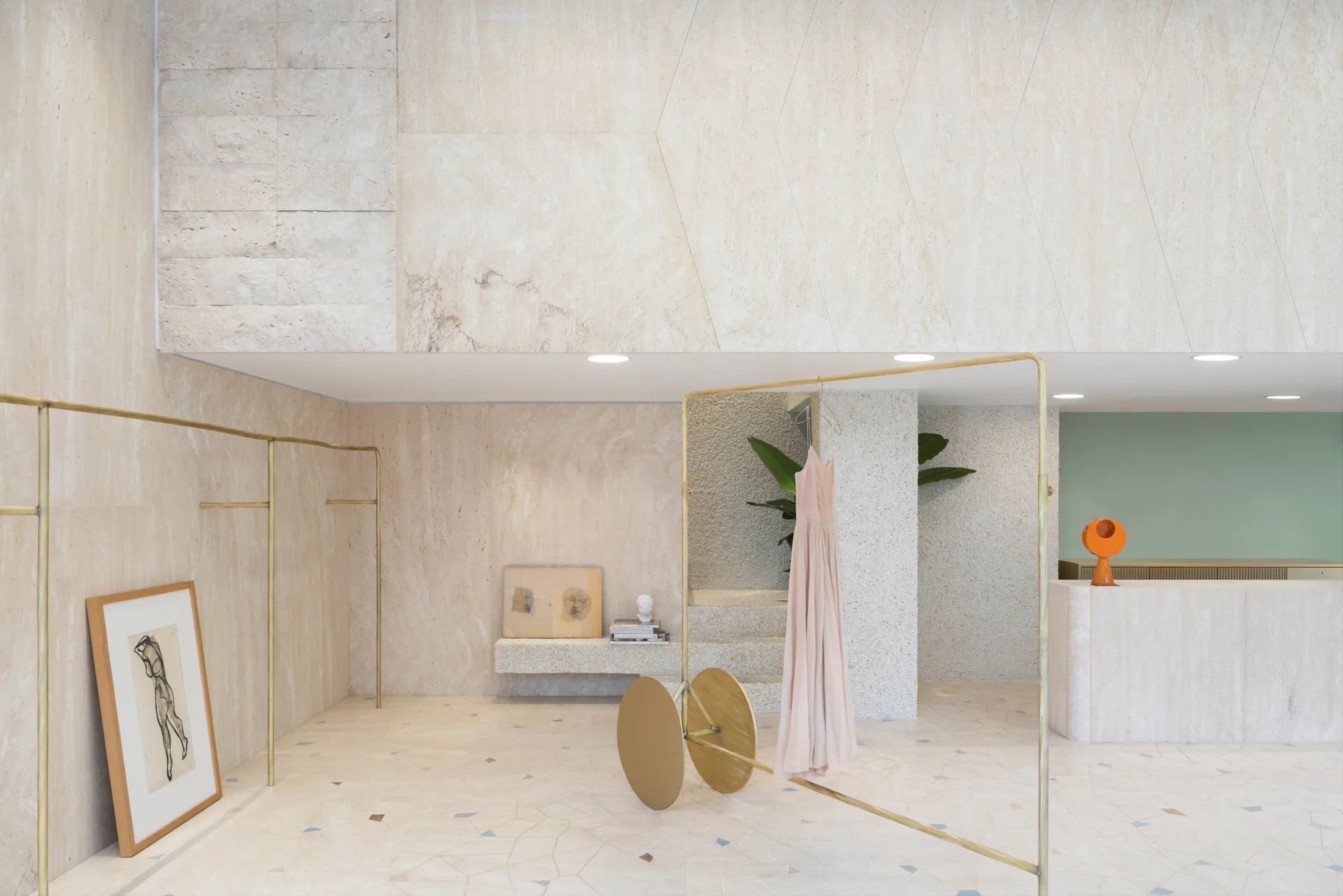 A minimalist retail space displays a pink dress on a gold metal rack against beige travertine stone walls and terrazzo-like flooring.