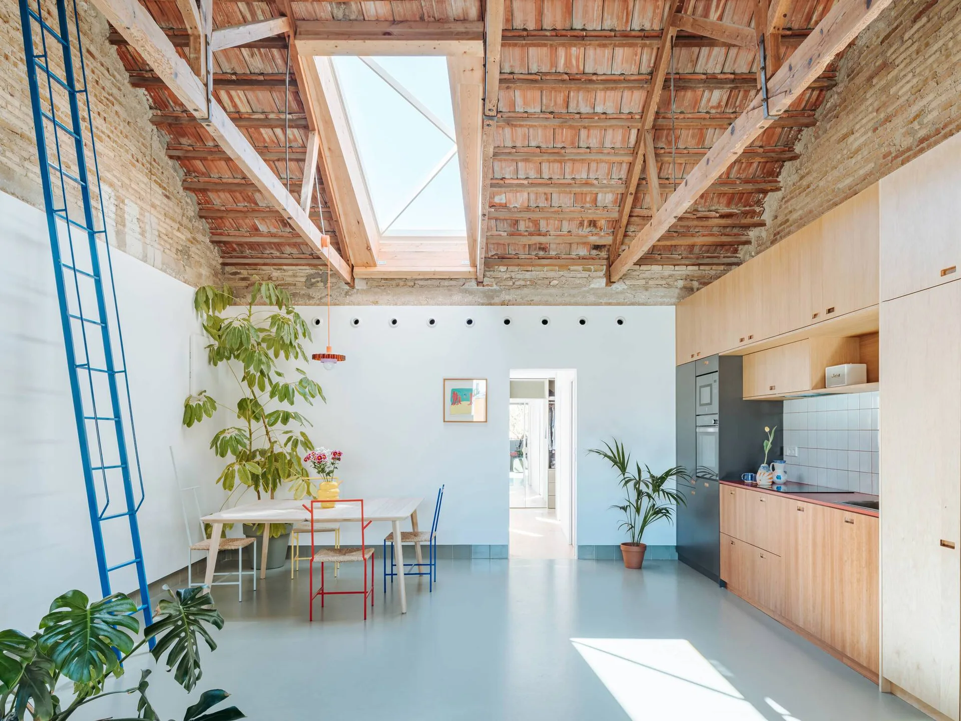 An open-concept kitchen and dining area features a pitched wood ceiling with a skylight, light wood cabinetry, colorful metal chairs, and a tall blue ladder.