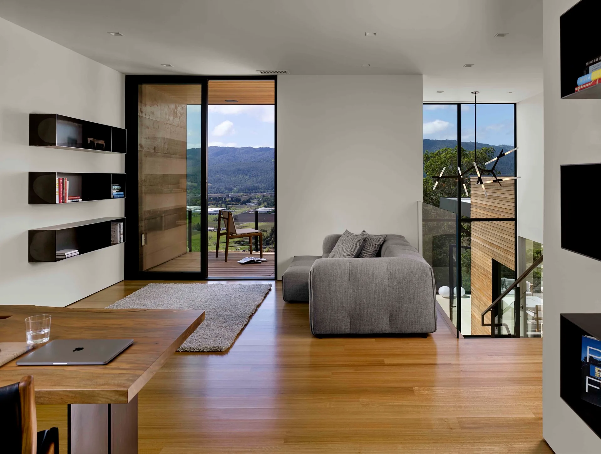 A modern living area featuring a grey sofa, a wooden desk in the foreground, black floating wall shelves, and glass doors opening to a scenic balcony.