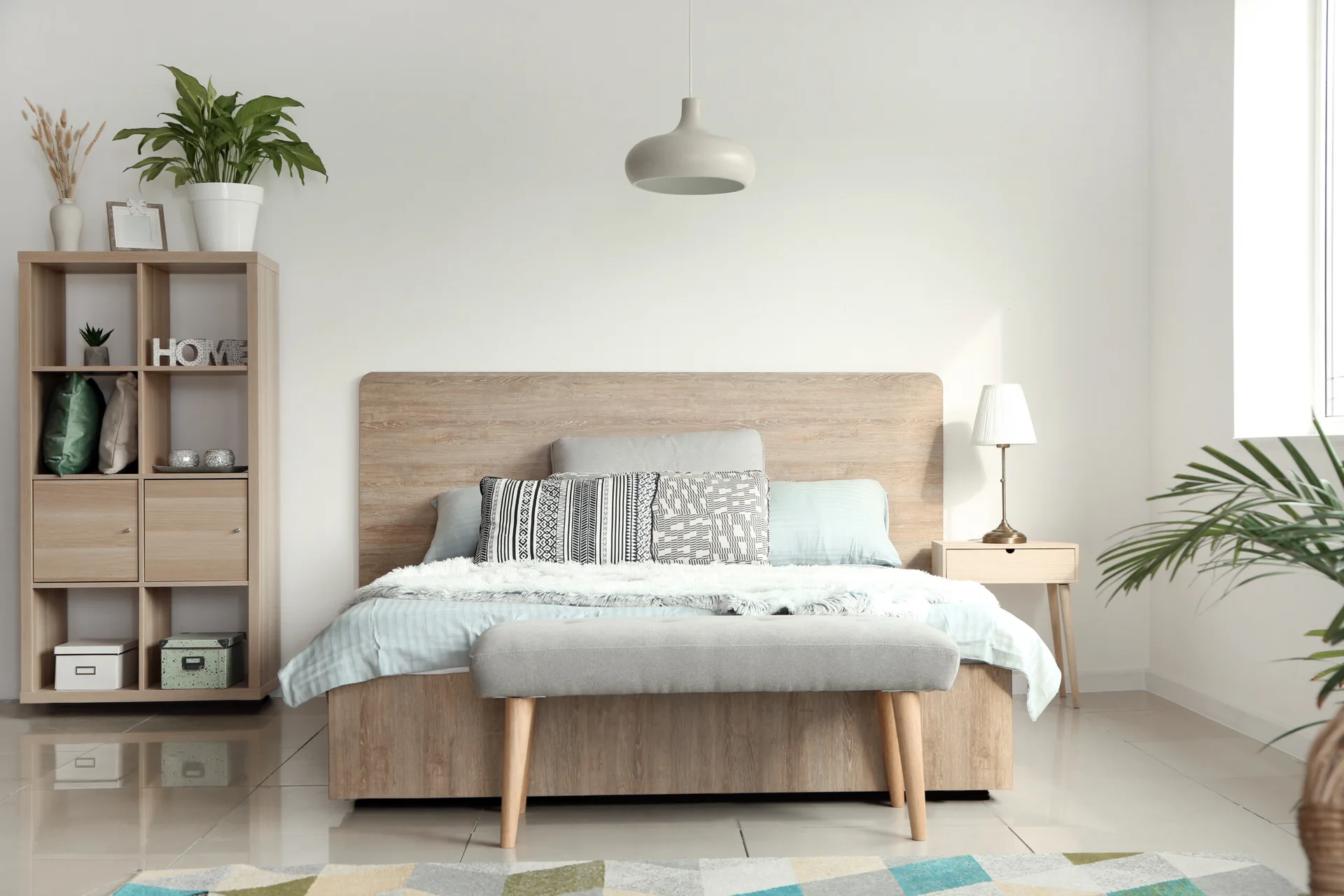 A bright bedroom featuring a light wood platform bed, an upholstered grey bench, and a matching tall bookshelf against white walls.