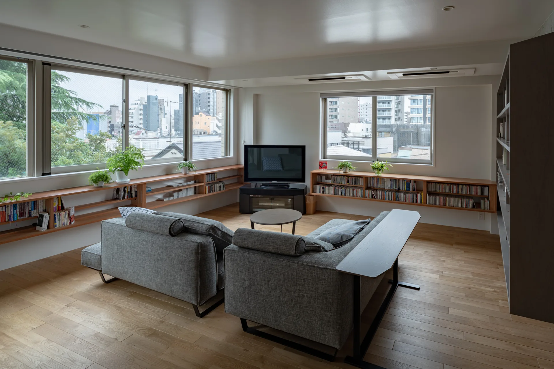 Two grey fabric sofas sit on light wood floors in an apartment living room featuring low built-in wooden bookshelves beneath large windows.
