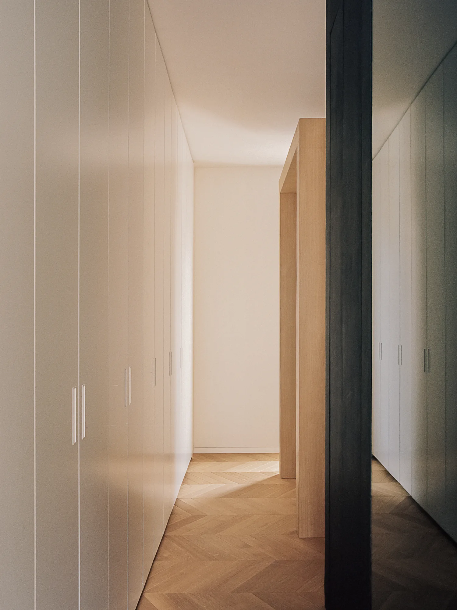 A narrow minimalist hallway features floor-to-ceiling white built-in wardrobes and a light oak herringbone patterned wood floor.